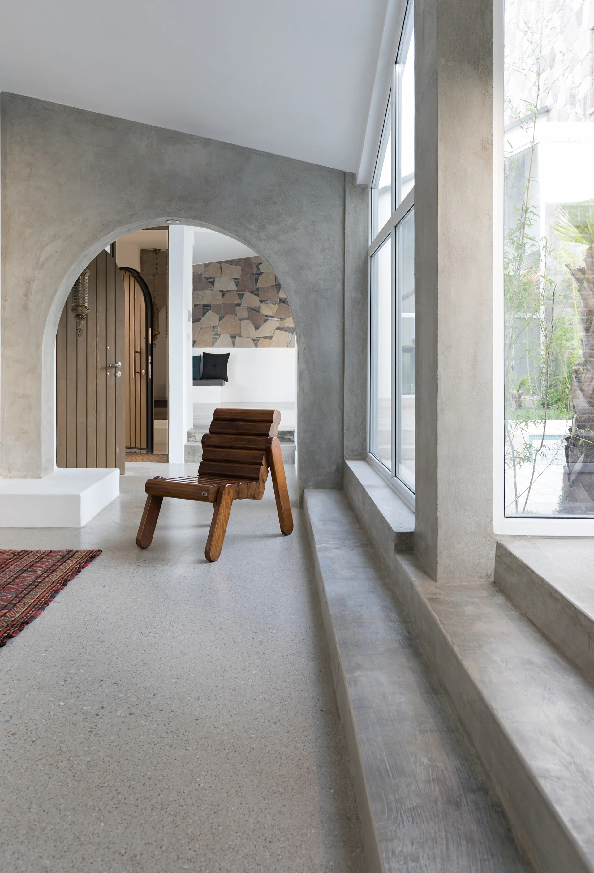 A slatted wooden chair sits near a raw concrete arched entryway in a minimalist room with large windows and a concrete floor.