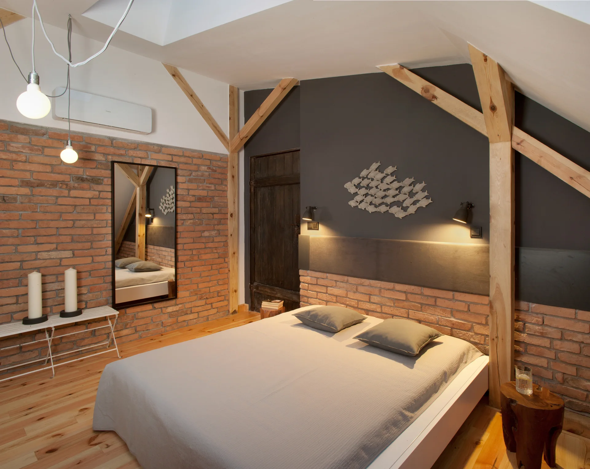 A rustic attic bedroom featuring a low-profile bed, an exposed brick wall, natural timber support beams, and a dark charcoal grey accent wall.