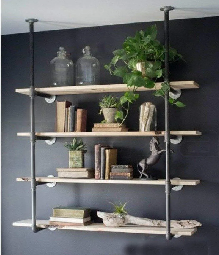 An industrial wall-mounted shelving unit with black iron pipes and light wood shelves is styled with books and plants against a dark grey wall.