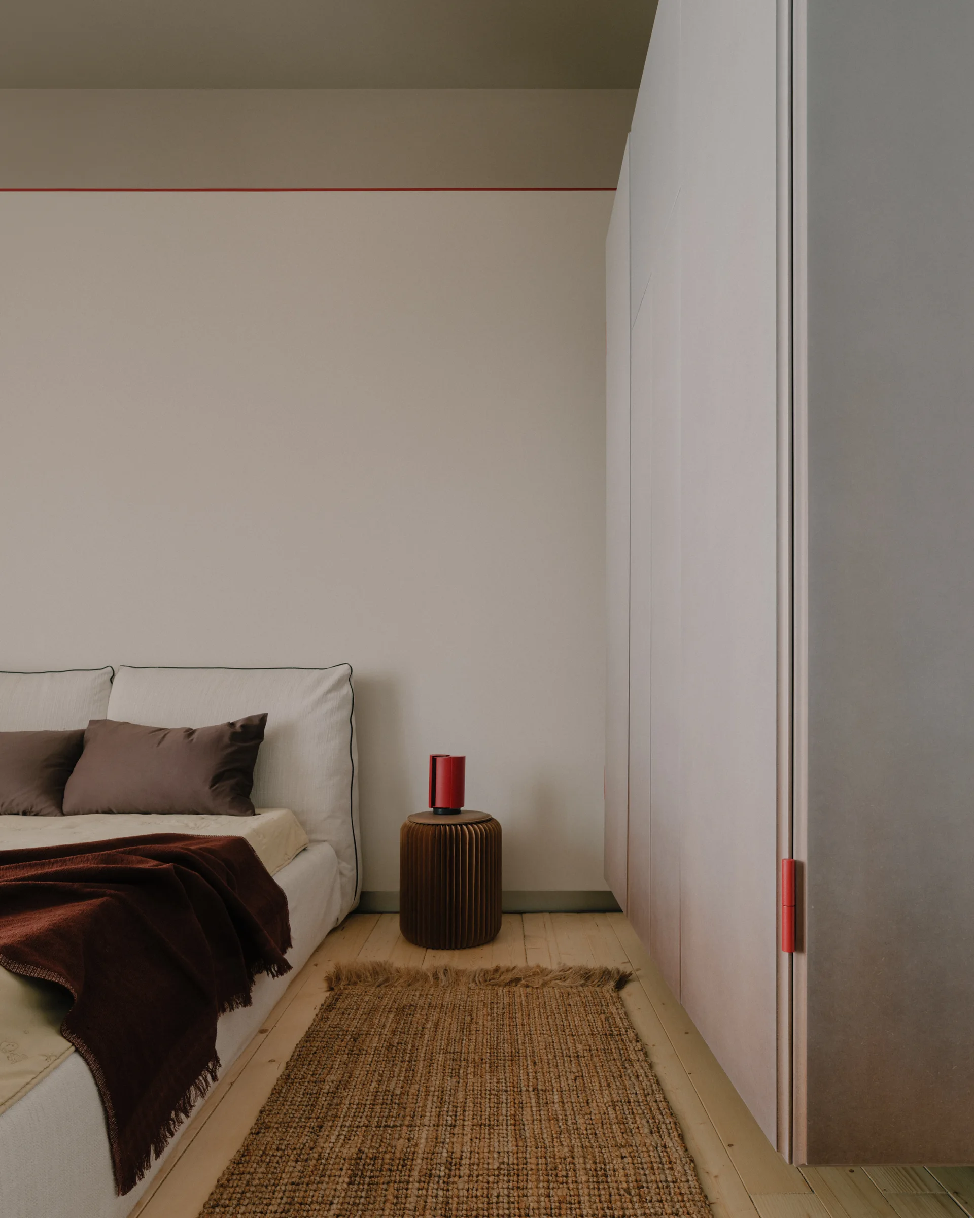 A minimalist bedroom features an upholstered bed with brown pillows, a ribbed cylindrical nightstand, and a tall wardrobe with red accents.