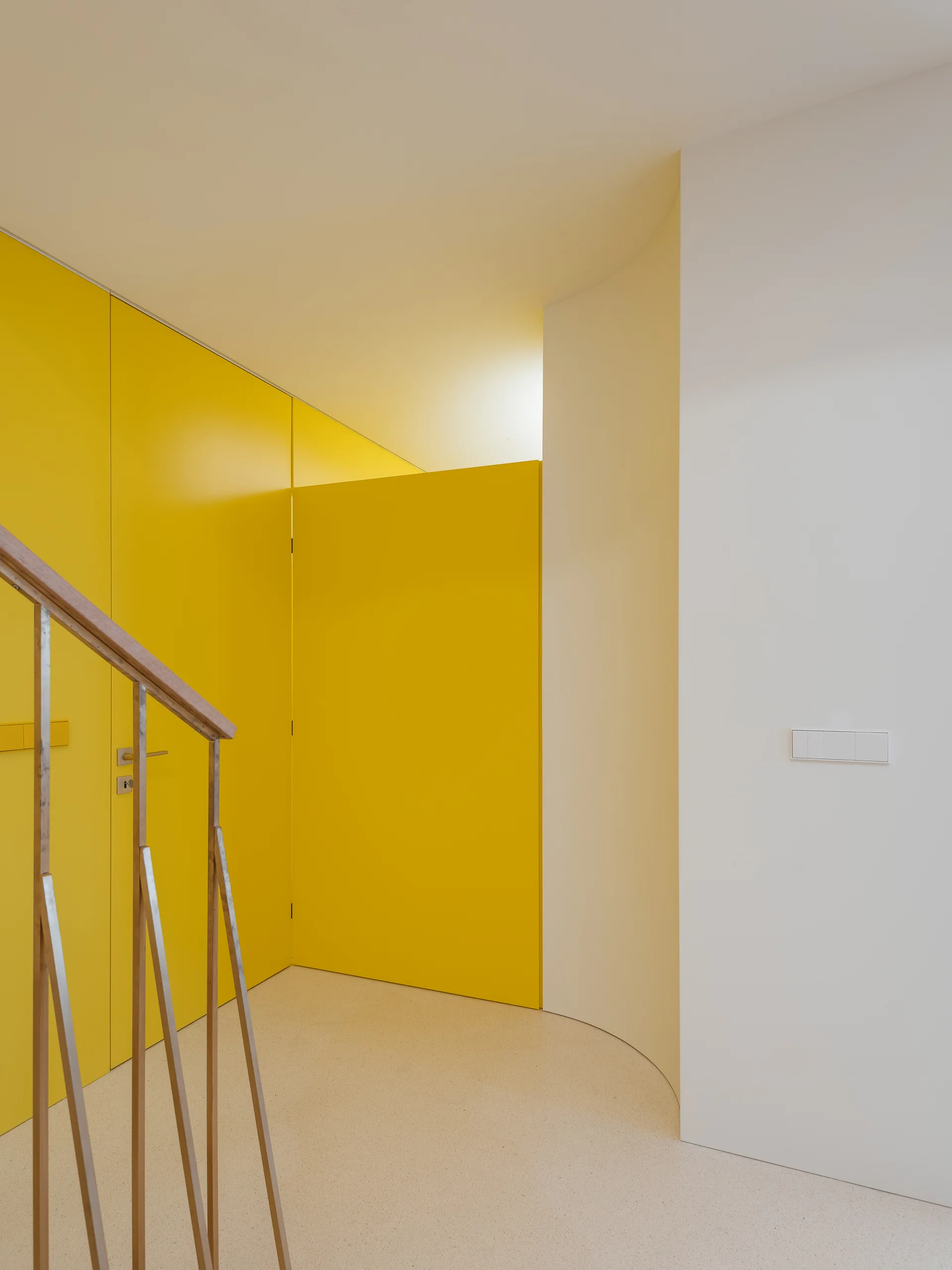 A modern entryway features a bright yellow floor-to-ceiling door system, a curved white wall, and a staircase railing with a wooden handrail.