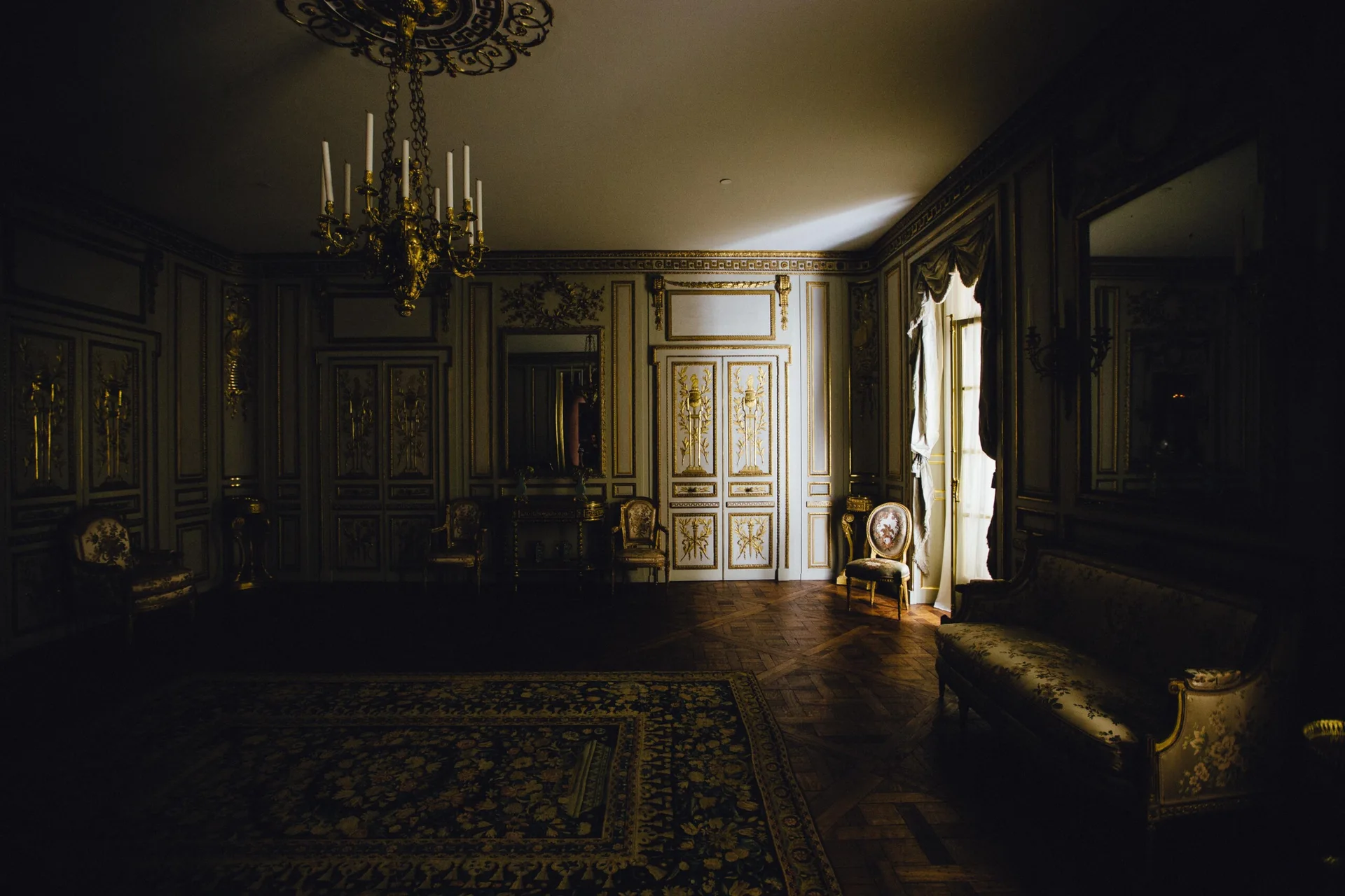 A dimly lit classical salon features ornate gilded wall paneling, a hanging gold chandelier, parquet wood flooring, and vintage upholstered furniture.