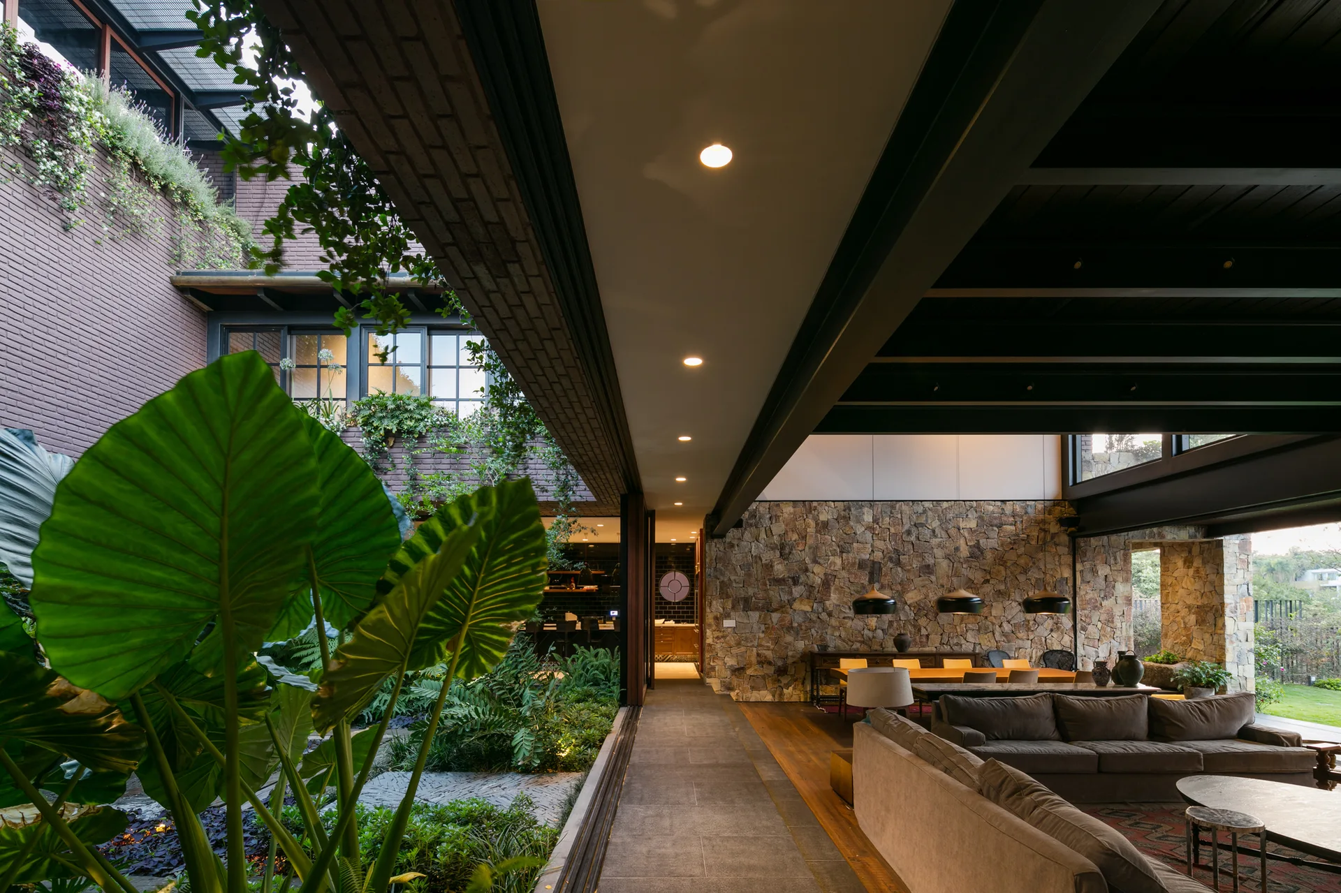 A modern living room with a grey sofa and rustic stone wall sits next to an open-air central courtyard filled with lush green plants.