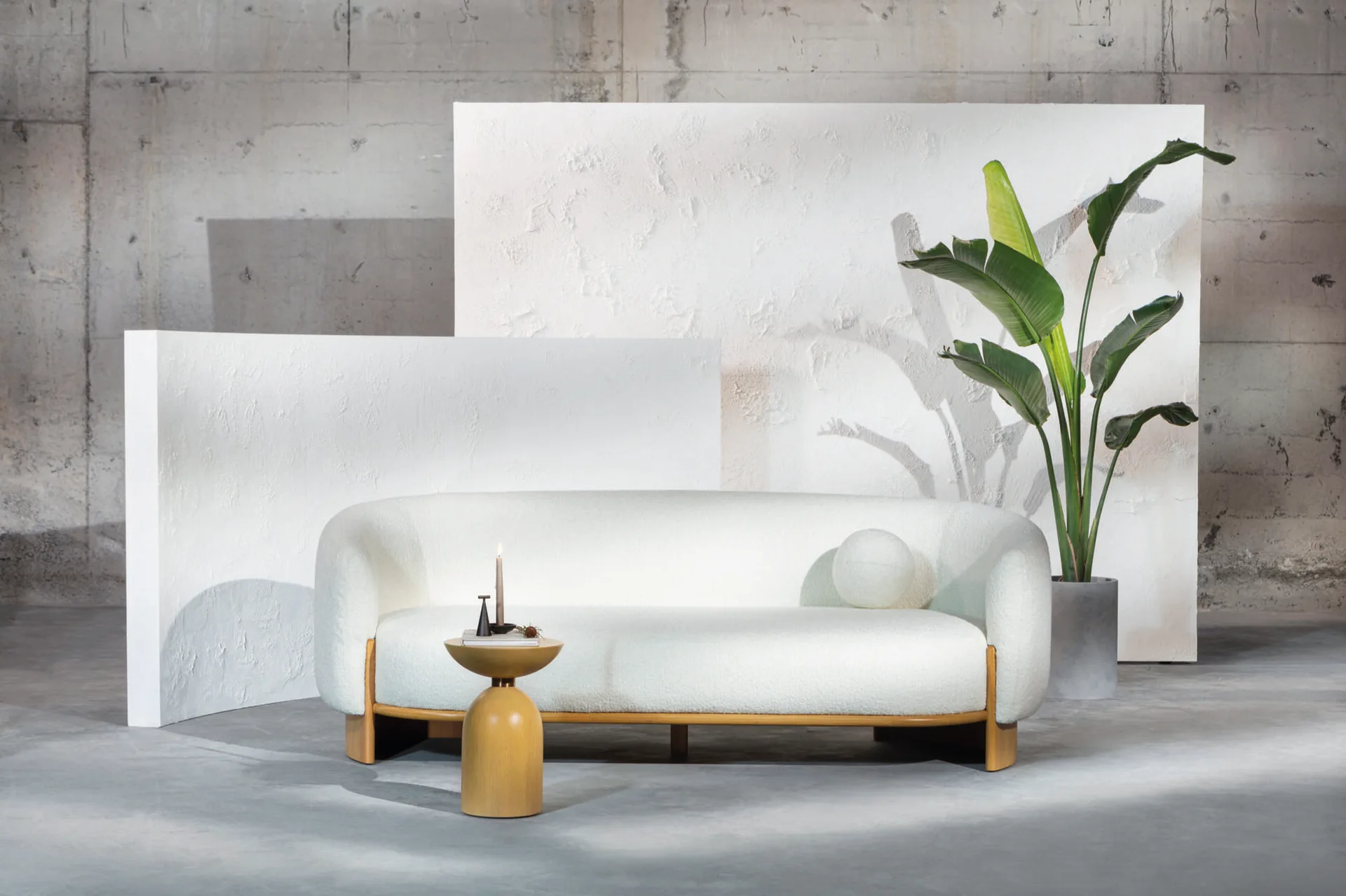 A curved white boucle sofa with a wood base sits in front of textured white screens next to a tall potted plant and gold side table.
