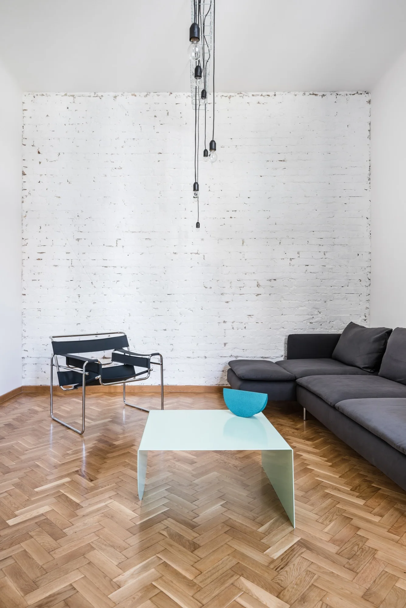 A minimalist living room features a dark gray sofa, a modern black accent chair, and a pale blue coffee table on a herringbone wood floor.