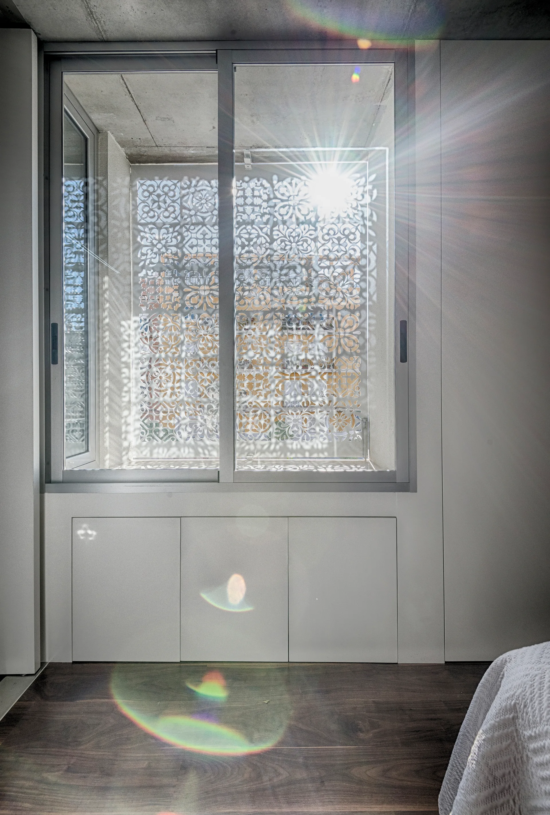 Sunlight streams through a decorative laser-cut screen outside a bedroom window, casting intricate shadows over white walls and built-in cabinets.
