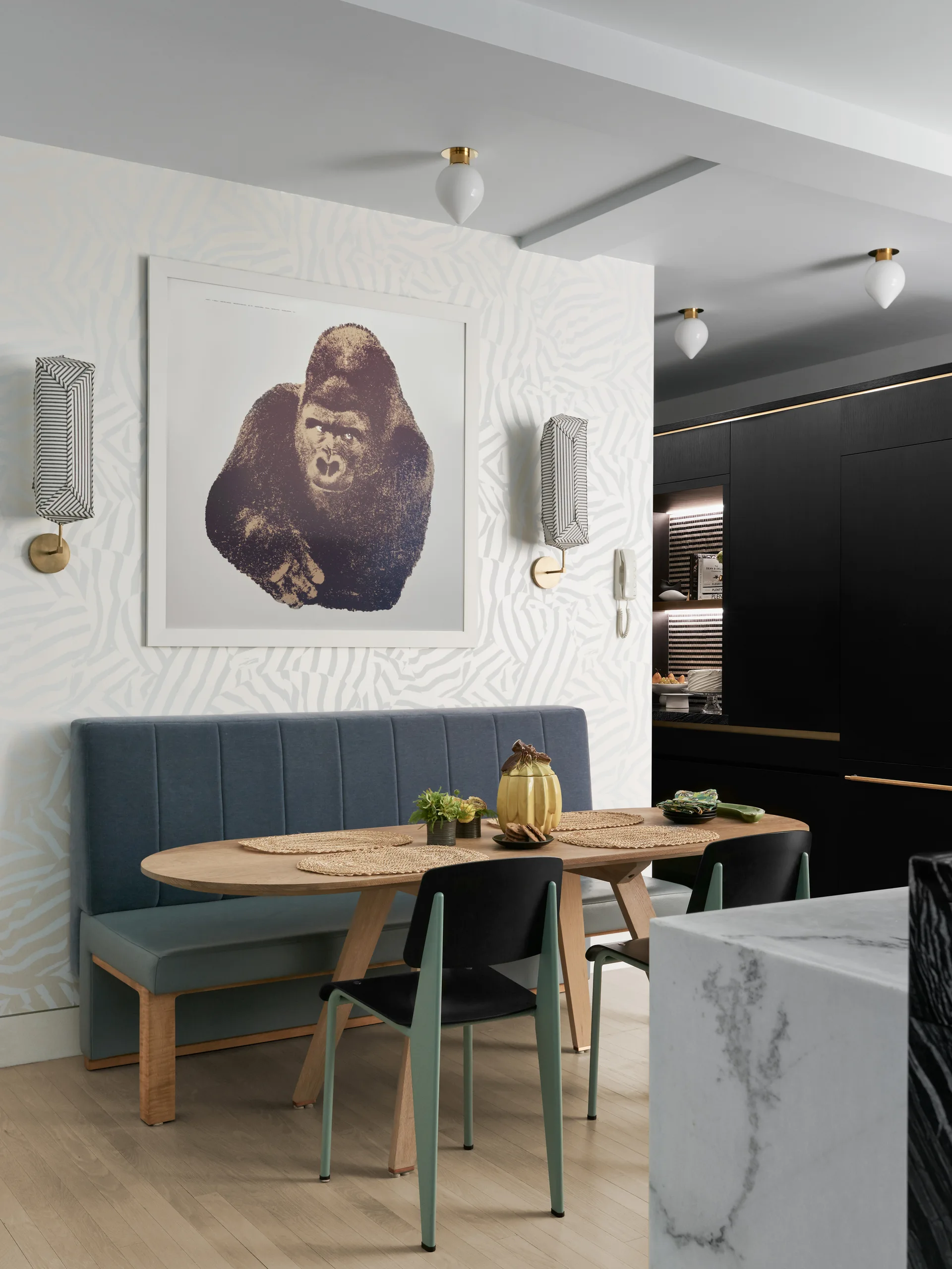 A modern dining nook features an oval wooden table, a blue upholstered banquette, and a framed gorilla print on zebra-patterned wallpaper.