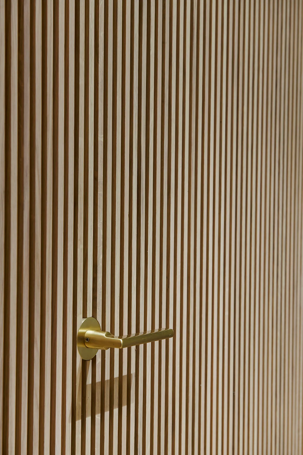 A close-up view of a minimalist satin brass lever door handle installed on a vertical light oak slatted wood accent wall.