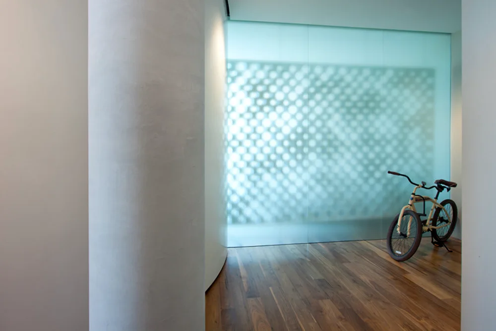 A minimalist hallway features warm hardwood floors, a curved white structural column, and a large frosted glass partition wall with a patterned shadow.