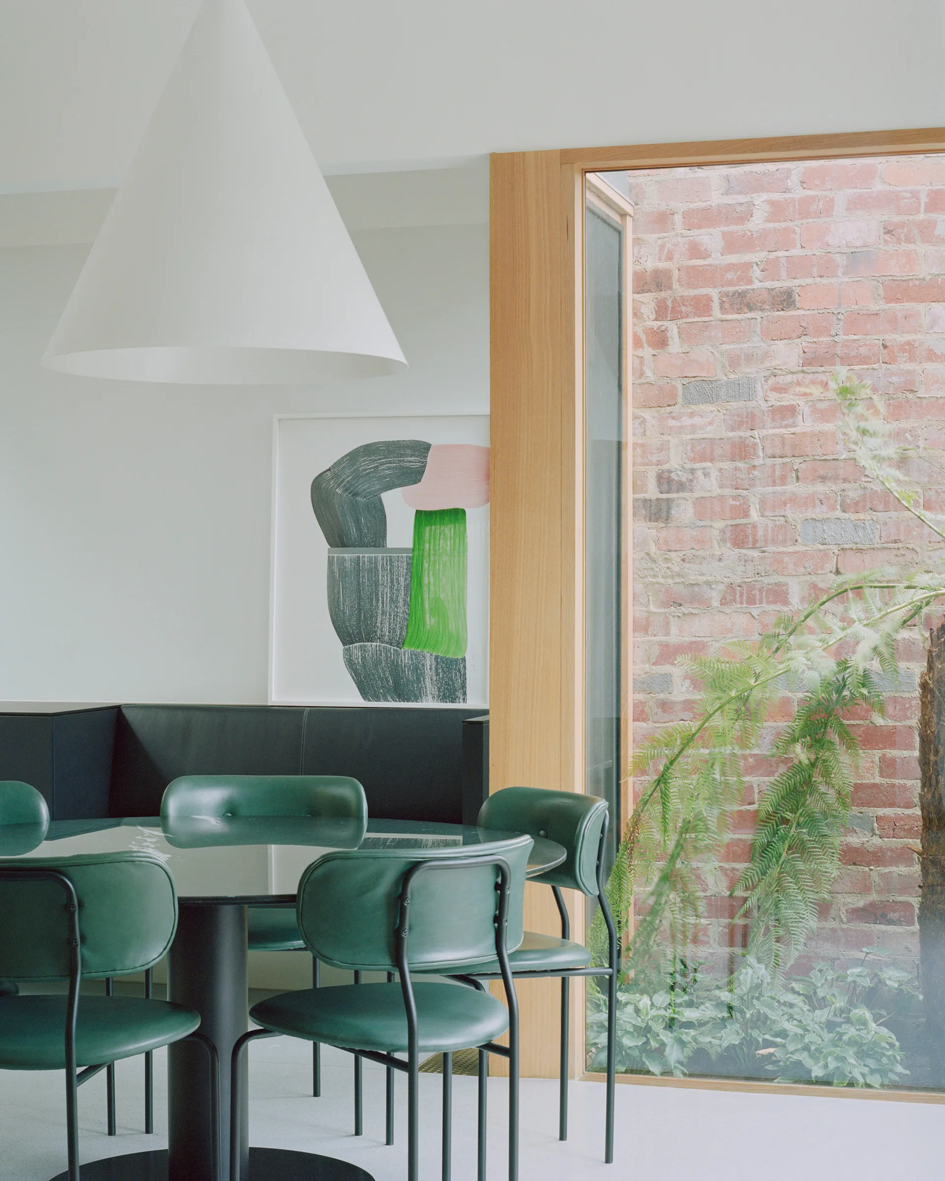 Green leather chairs surround a black table beneath a large white cone pendant light next to a window overlooking an outdoor brick wall.