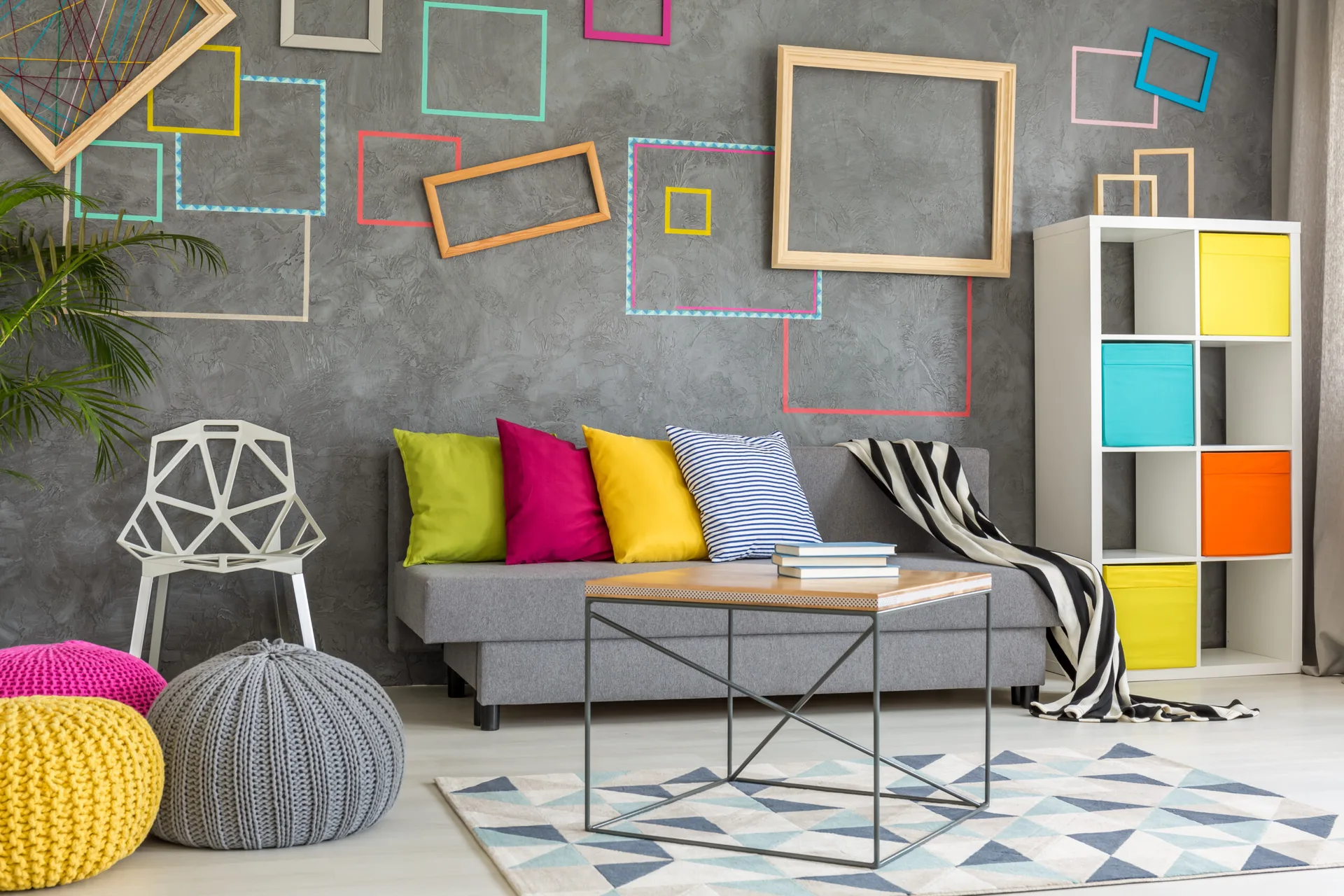 A grey sofa with colorful pillows sits in front of a textured grey wall decorated with empty frames next to a white cube shelf.