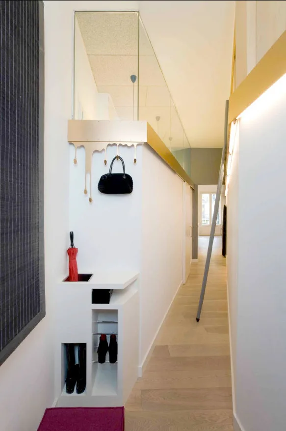 A modern white hallway features custom built-in storage, a playful gold drip-effect coat hook holding a handbag, and a slanted metal ladder.
