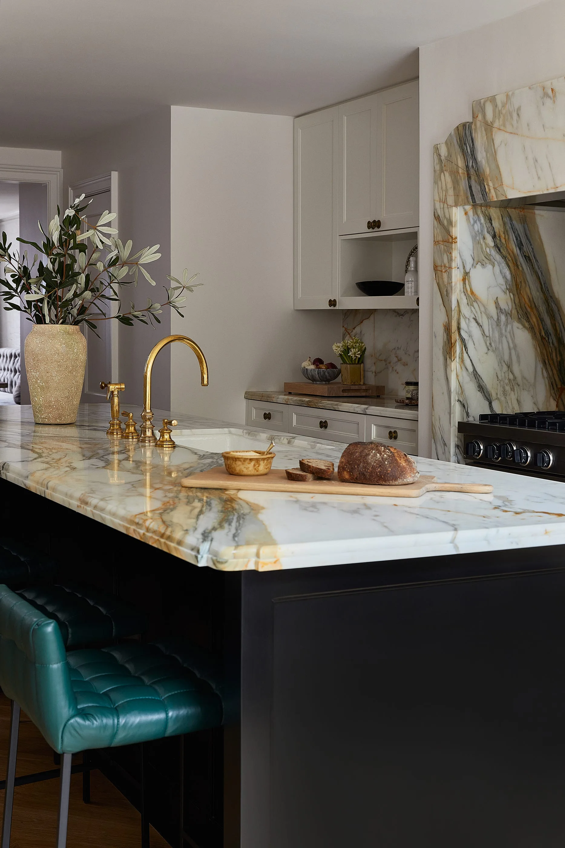 A kitchen island features a heavily veined white marble countertop, a brass faucet, and tufted emerald green leather bar stools.
