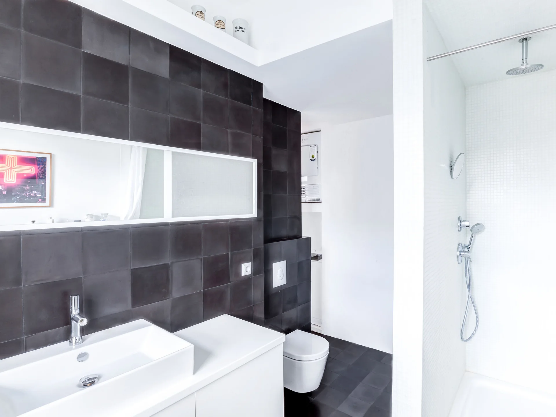 A minimalist bathroom features a wall covered in matte black square tiles, a white floating vanity with a chrome faucet, and a wall-mounted toilet.