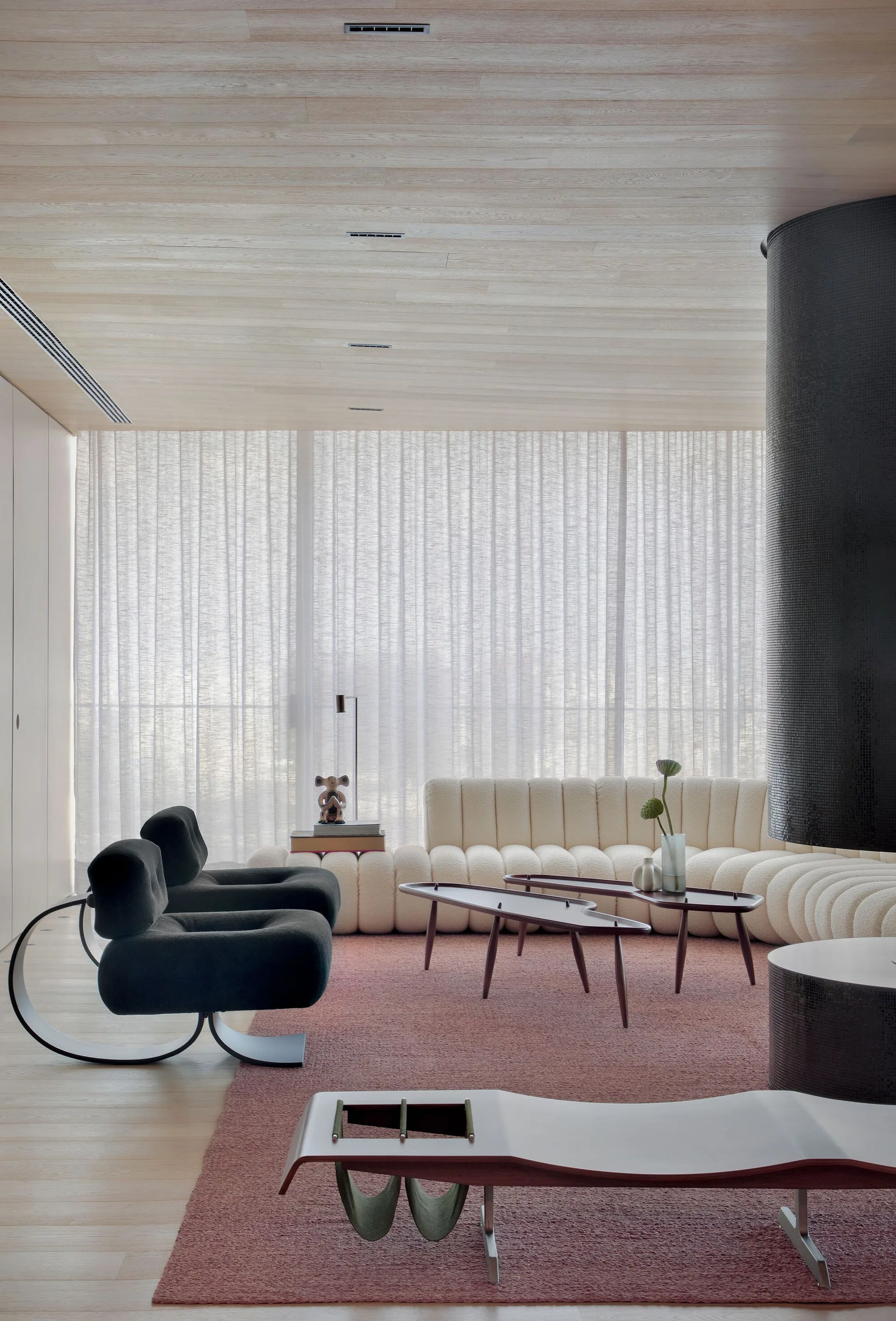 A modern living room features a cream channel-tufted sofa, two black modern lounge chairs, and a muted pink rug under a light wood plank ceiling.