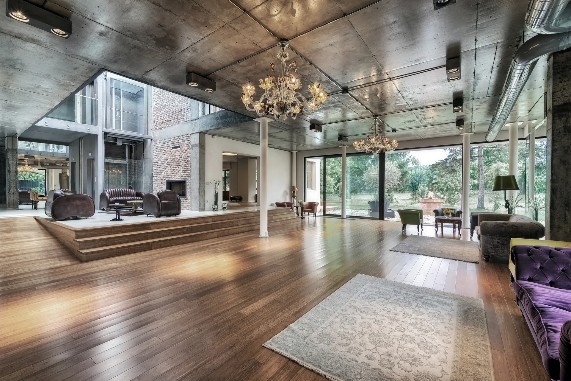 An industrial living room featuring a raw concrete ceiling, a raised wooden platform with leather chairs, and large windows.