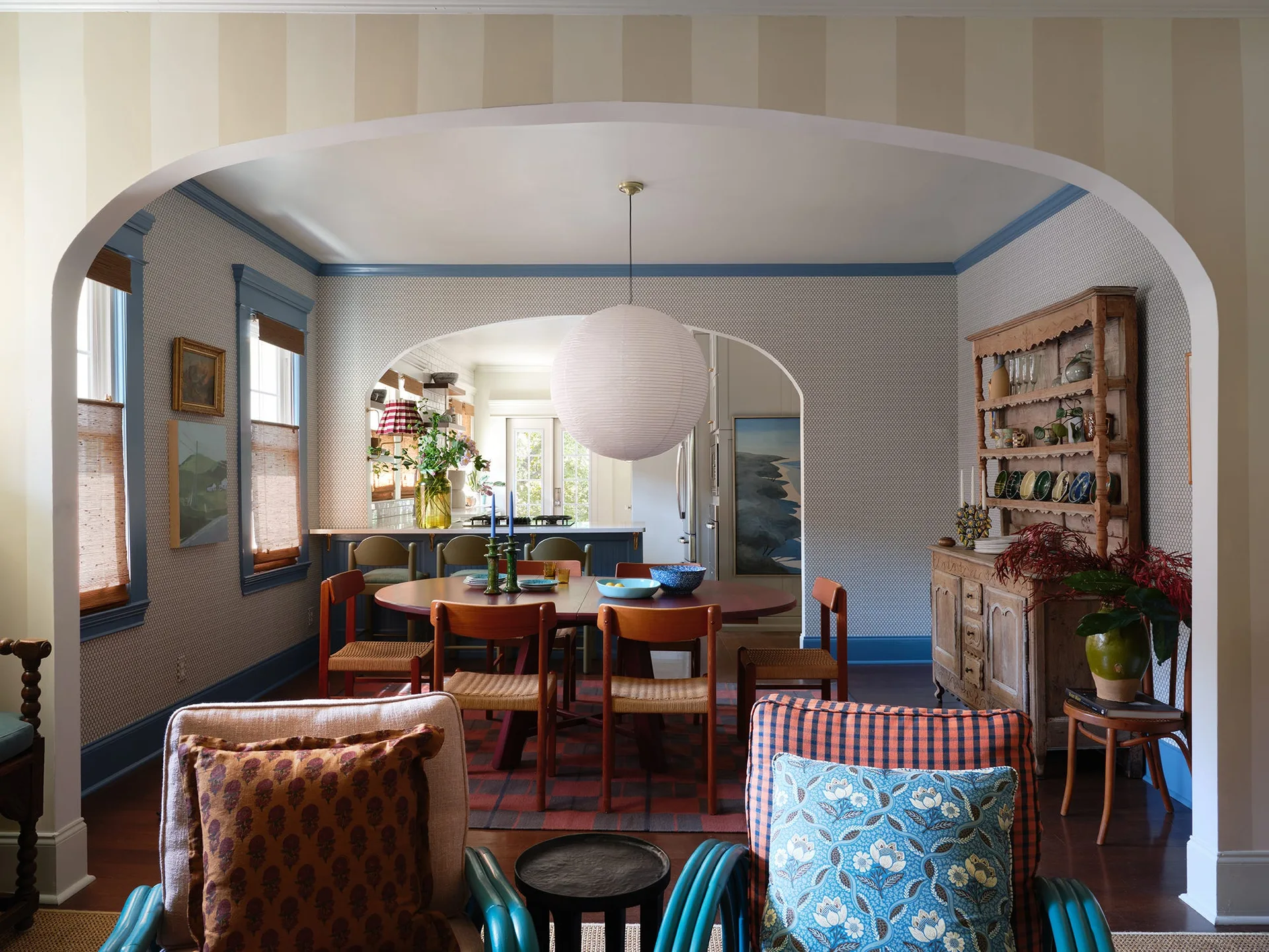 An eclectic dining room with a wooden table, blue trim, patterned wallpaper, and a paper globe pendant light, seen through a striped archway.