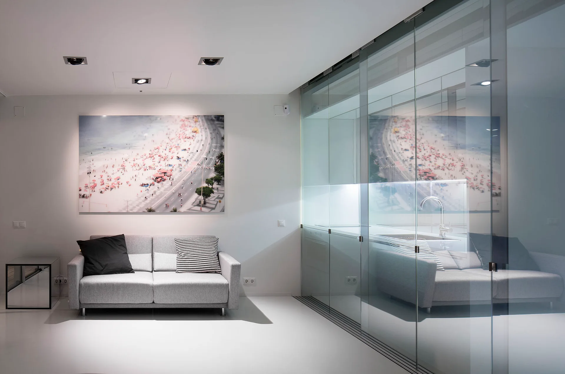 A grey sofa with pillows sits against a white wall next to a large beach photograph and a sliding glass partition.