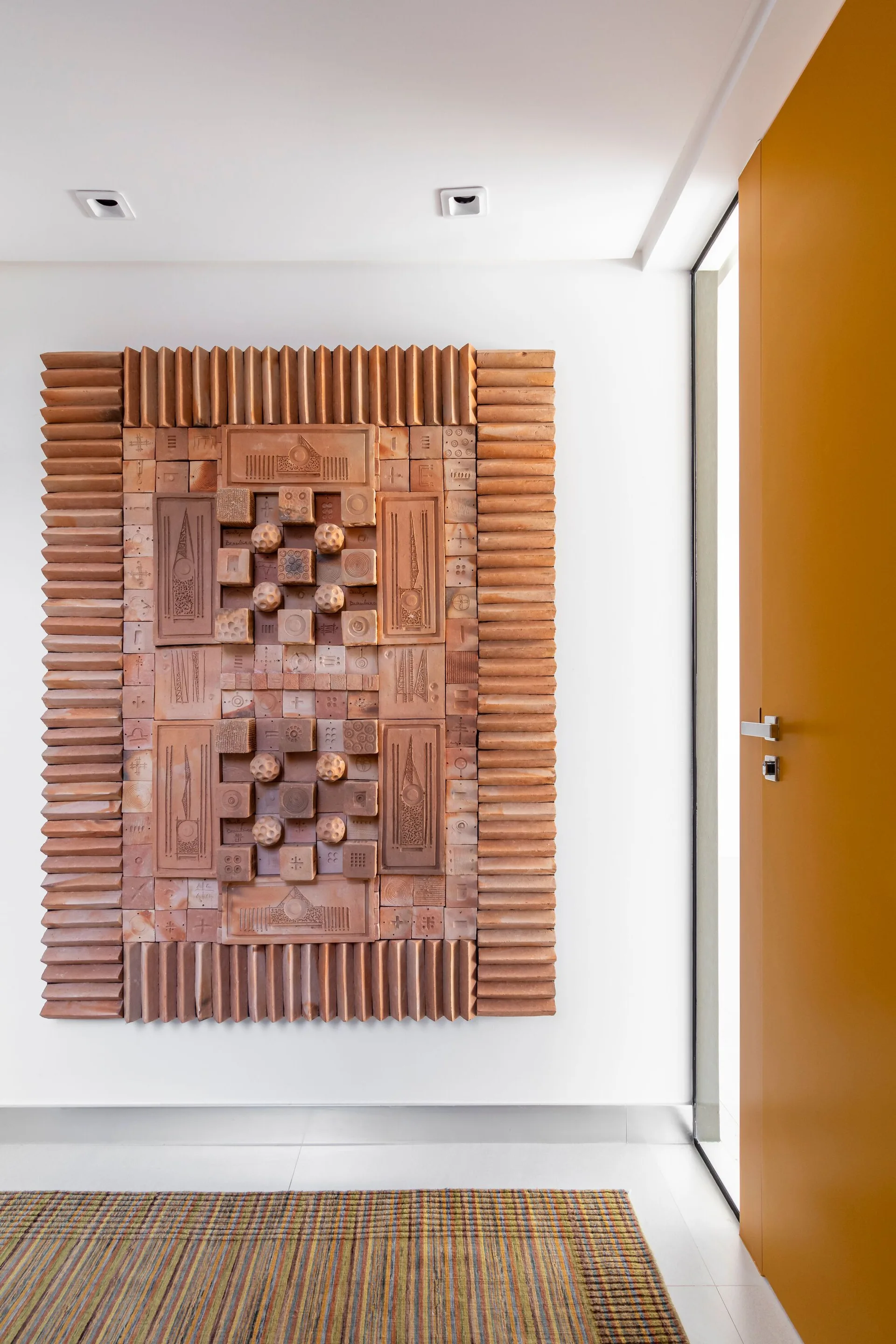 A large geometric terracotta clay relief sculpture hangs on a white entryway wall next to a bright yellow door.