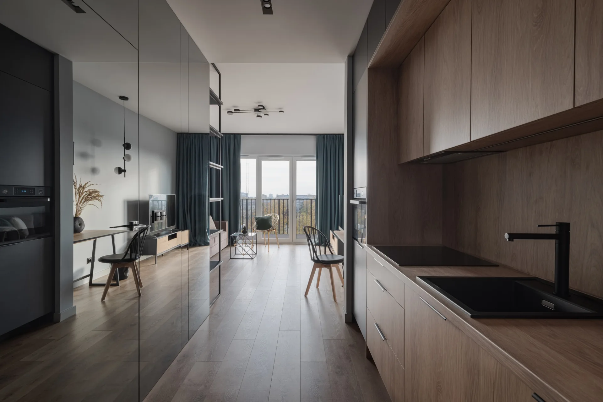 A modern narrow apartment corridor with wood-toned kitchen cabinets on one side and a floor-to-ceiling mirrored wall reflecting the adjacent living room.