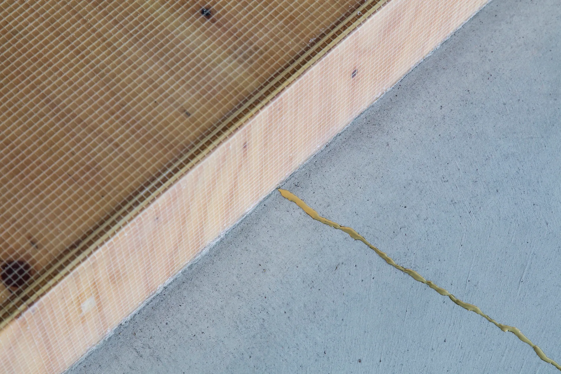 A close-up of a concrete floor with a yellow sealant seam next to a wood platform covered in wire mesh.