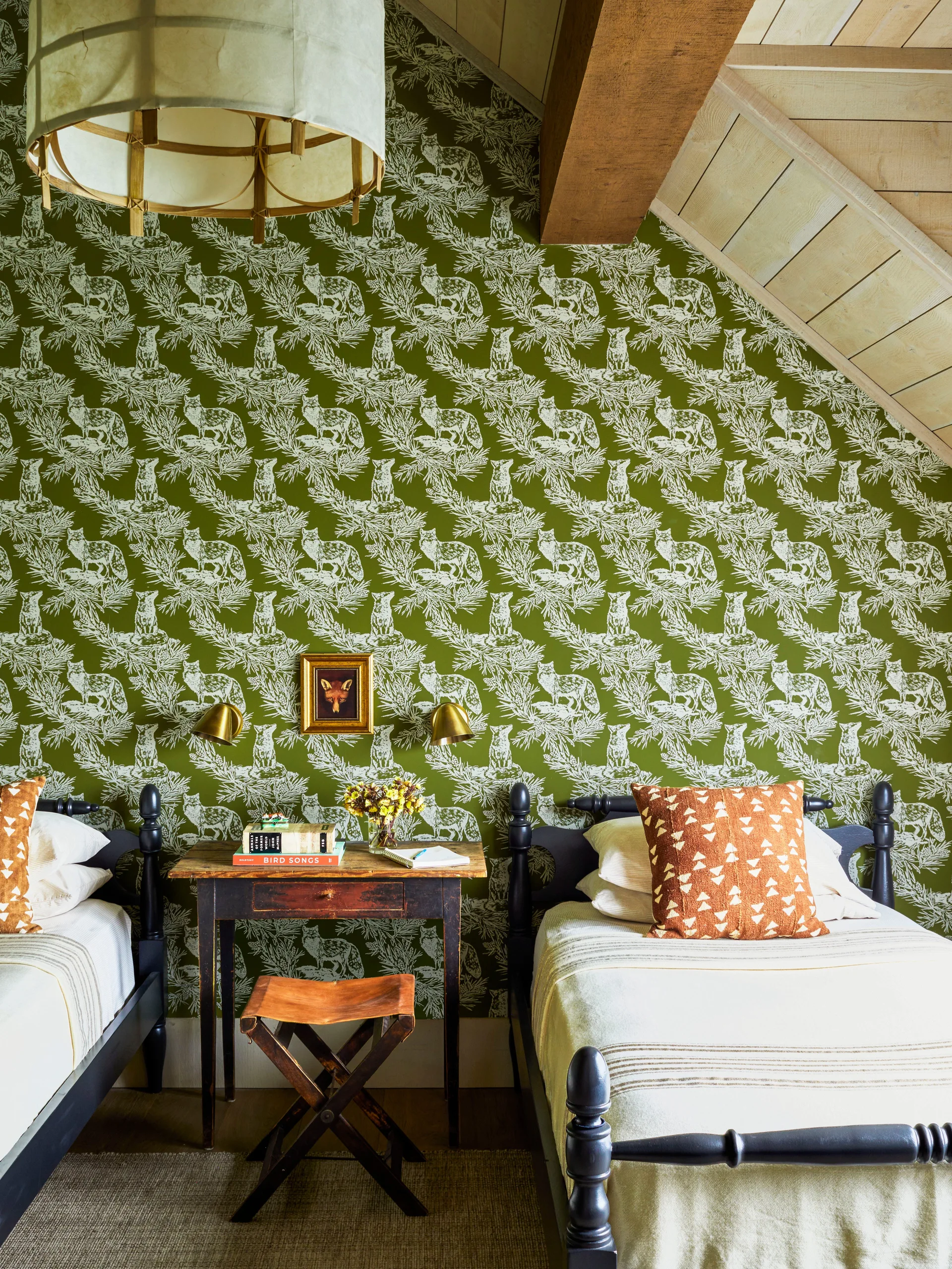 Two dark wooden twin beds flank a rustic nightstand against green fox-patterned wallpaper under a sloped, wood-paneled ceiling.