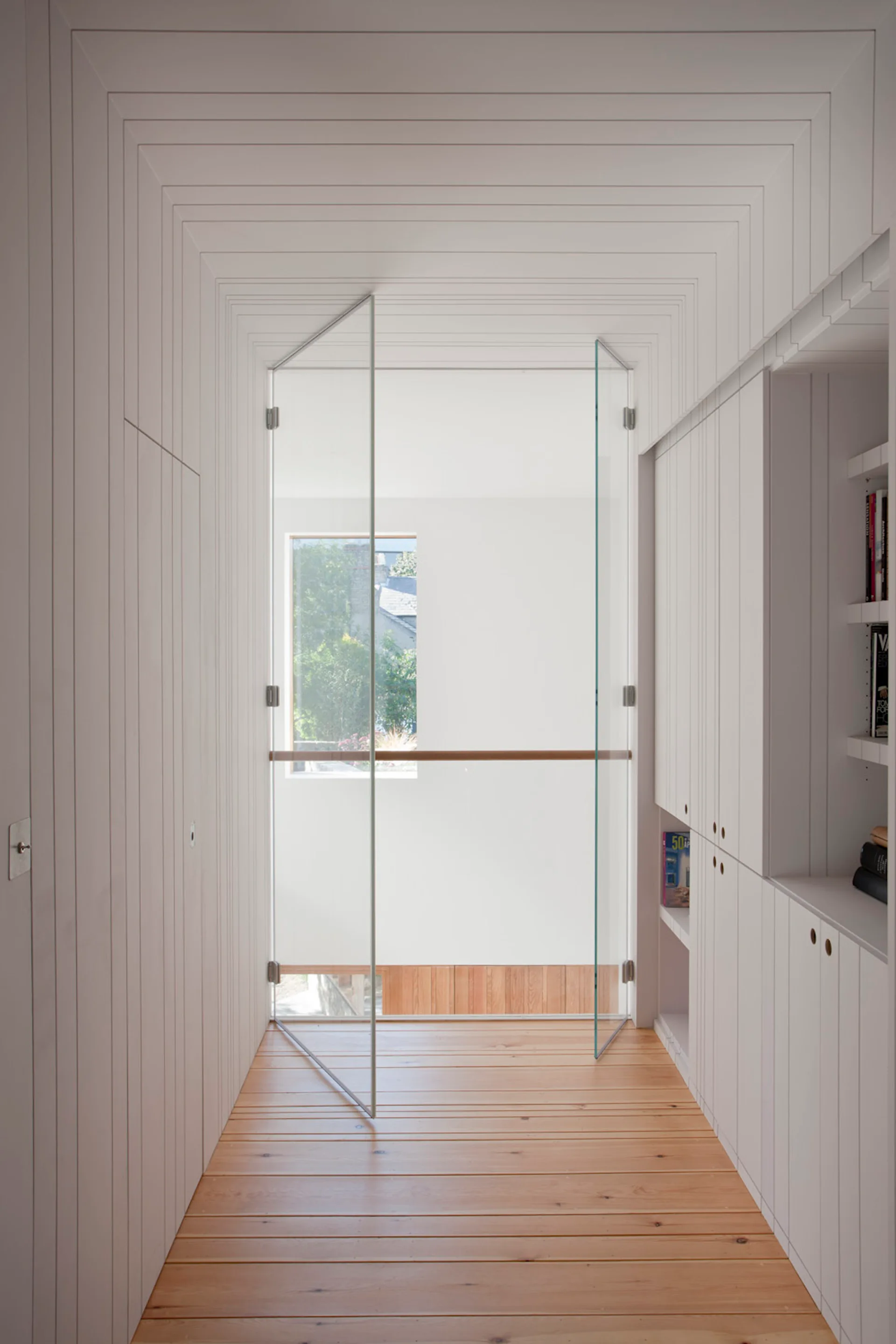A narrow hallway features white vertical wood-paneled walls and ceiling, natural wood floors, integrated white cabinets, and a frameless glass door.