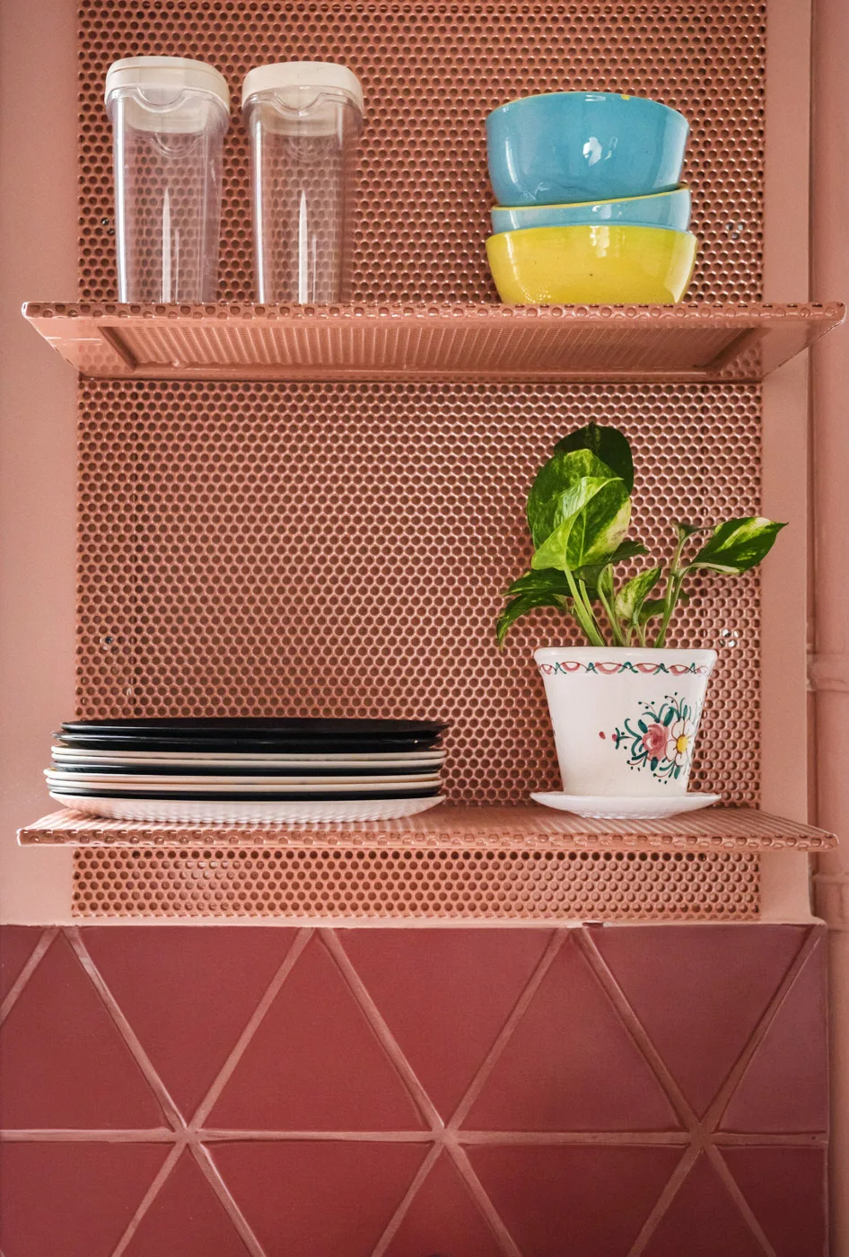 Two pink perforated metal shelves mounted on a matching back panel display kitchenware and a small potted plant above geometric triangle tiles.