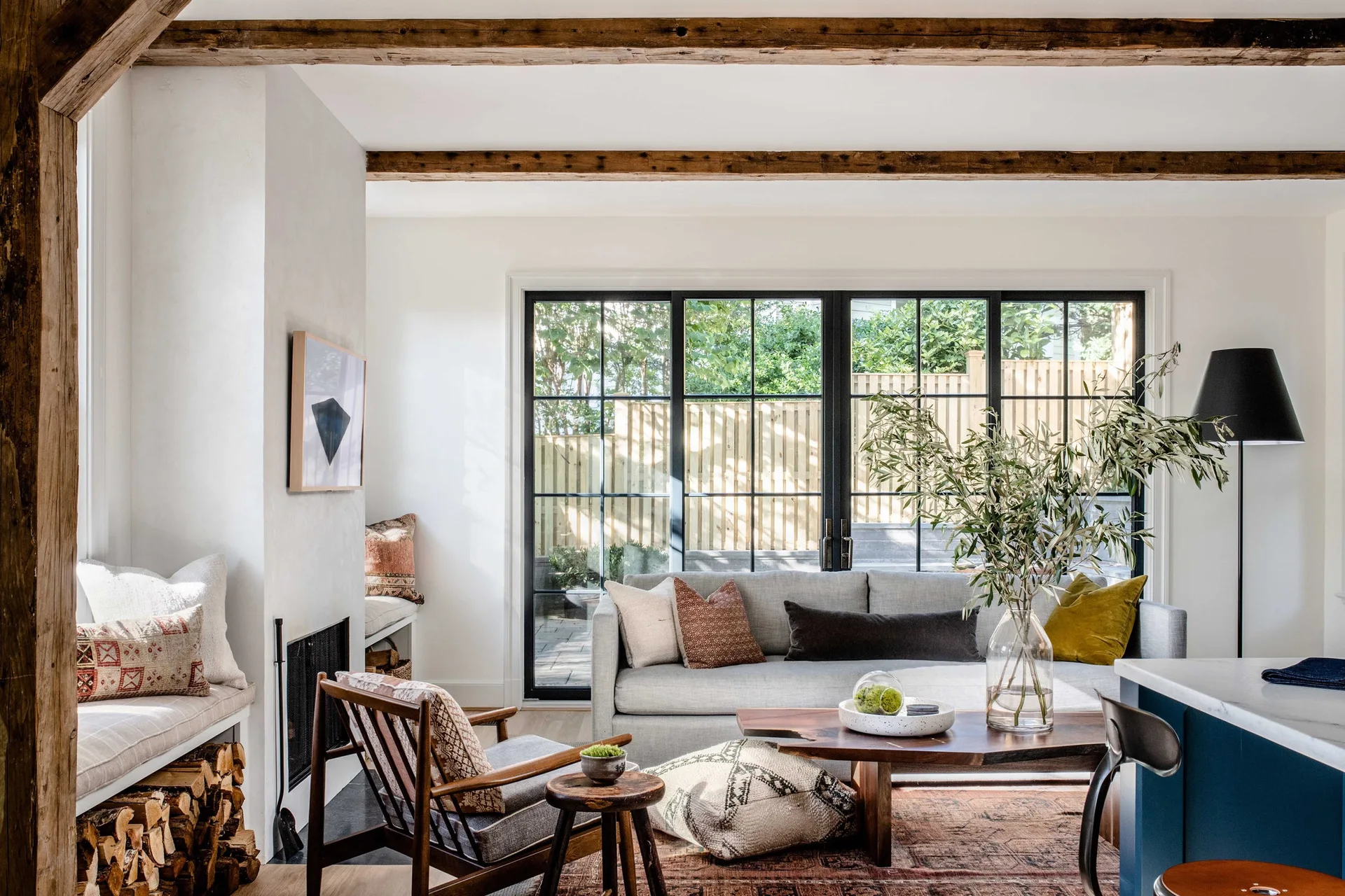 A bright living room features exposed wood ceiling beams, a grey sofa, a live-edge coffee table, and large black-framed glass doors leading outside.