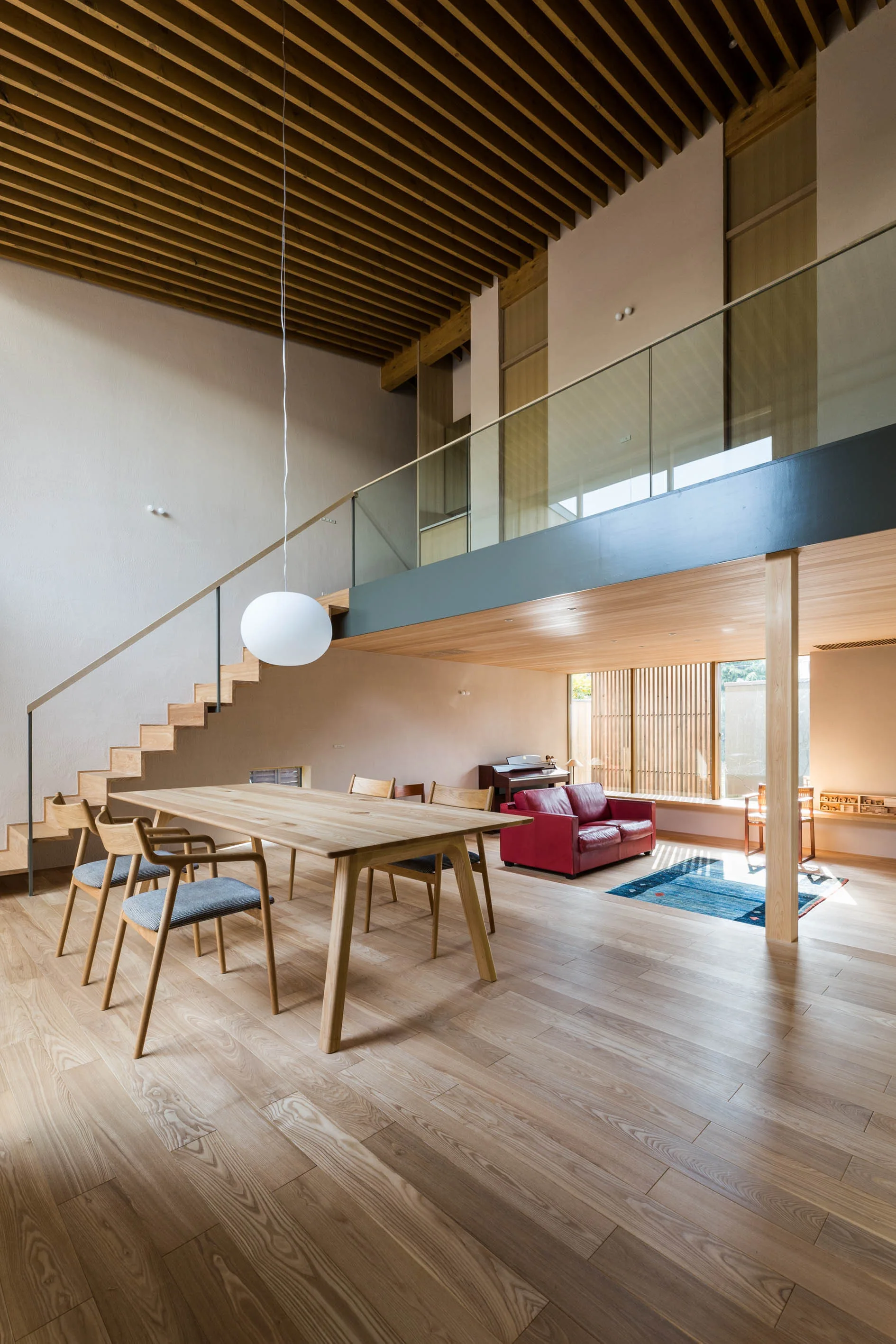 An open dining area features a light wood table, a floating staircase, an overhead mezzanine with a glass railing, and exposed ceiling beams.