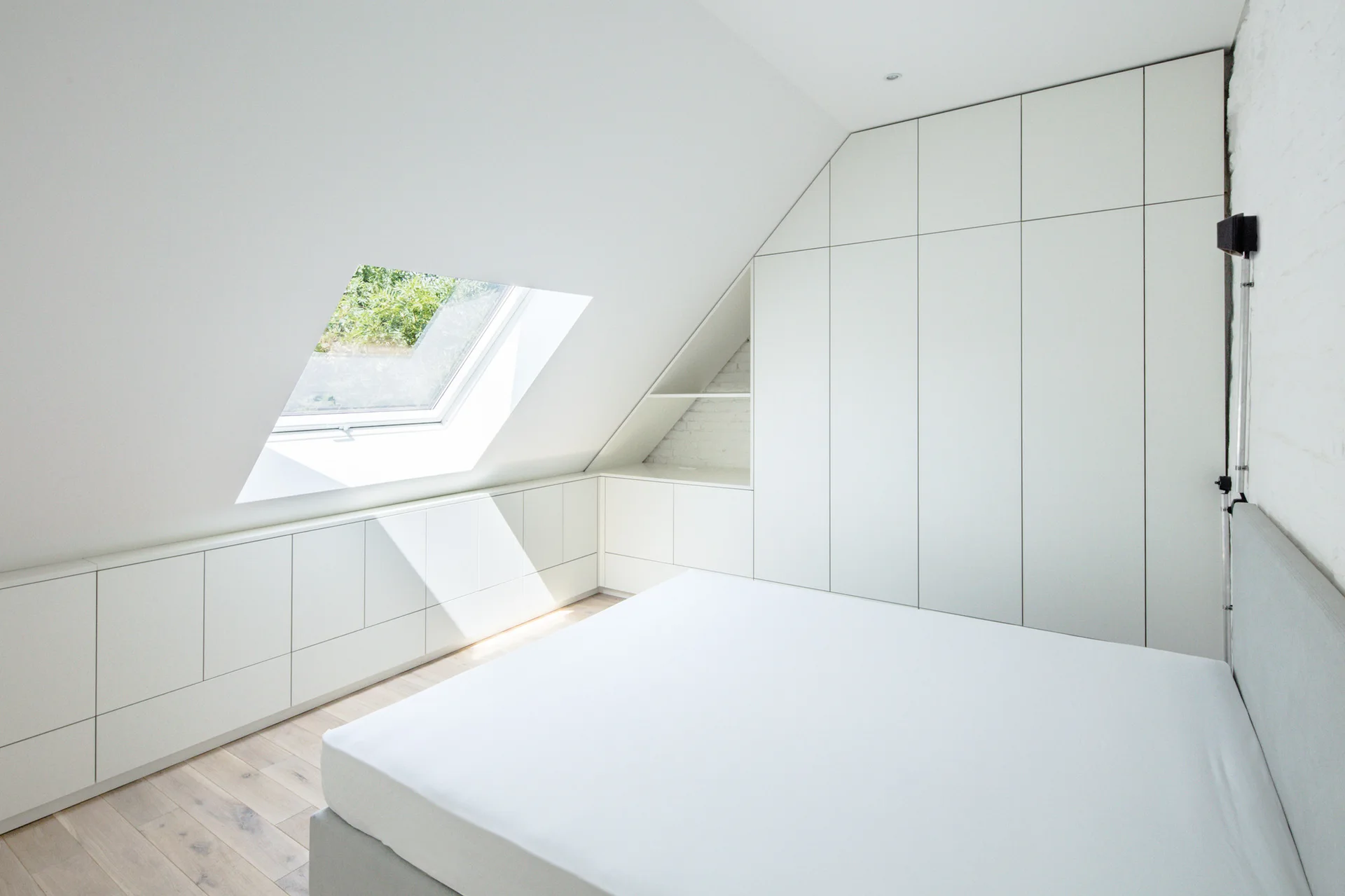 A minimalist attic bedroom featuring a bed, light wood flooring, a skylight, and flat-front white storage cabinetry tailored to the angled ceiling.
