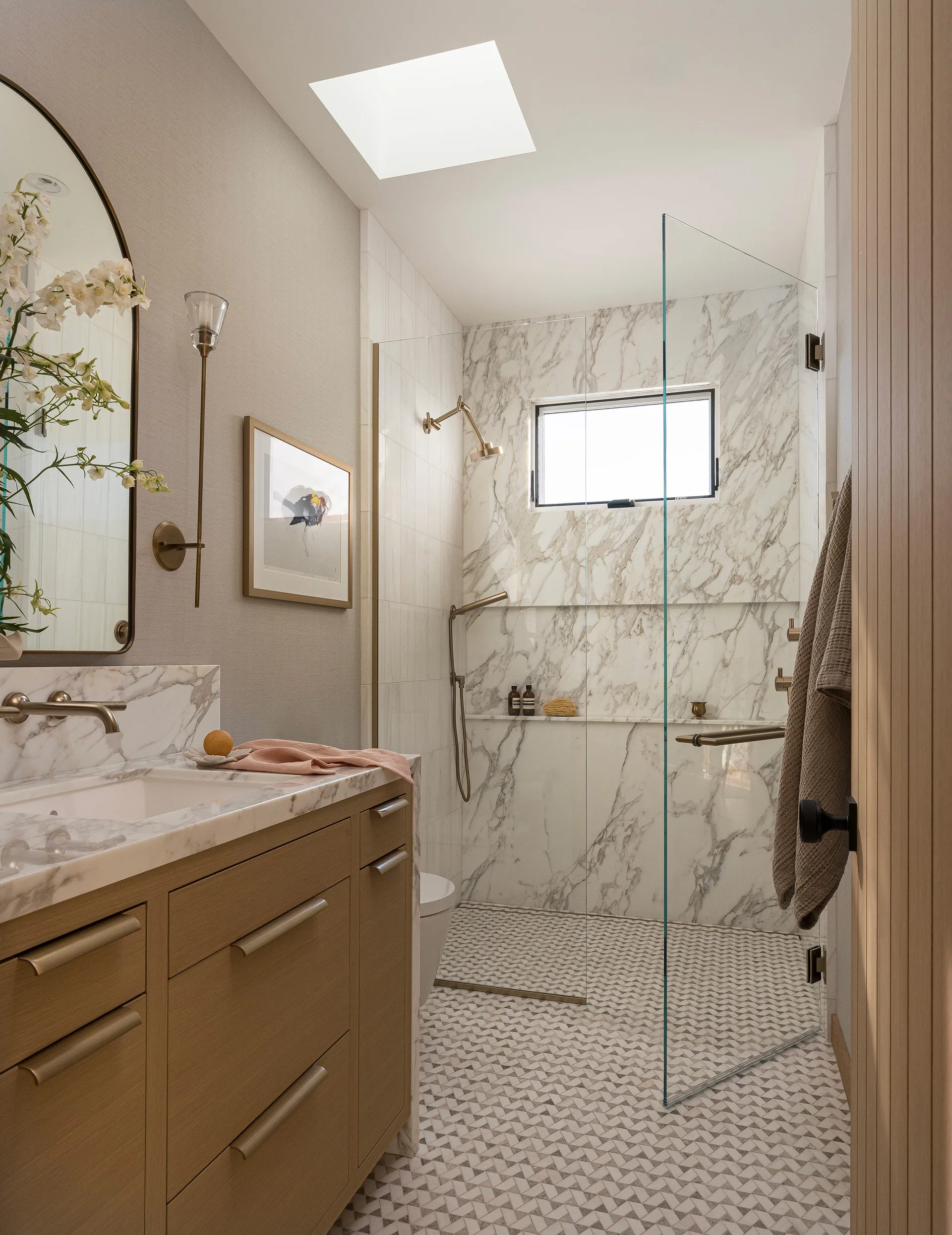 A modern bathroom with a light wood vanity, arched mirror, geometric tiled floor, and a glass walk-in shower with marble slab walls and a skylight.