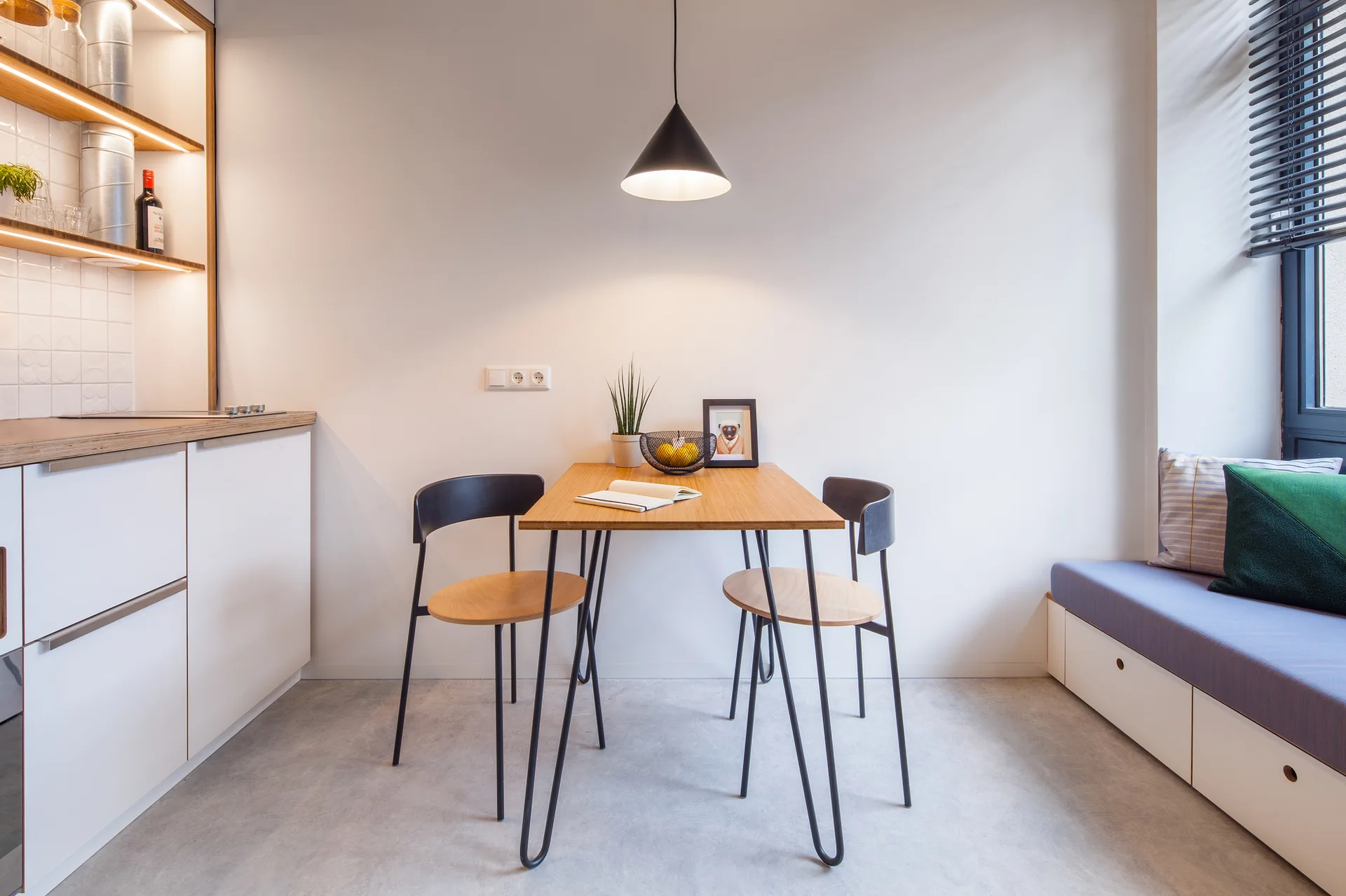 A small wooden dining table with hairpin legs stands between modern white kitchen cabinets and a built-in window bench with blue cushions.