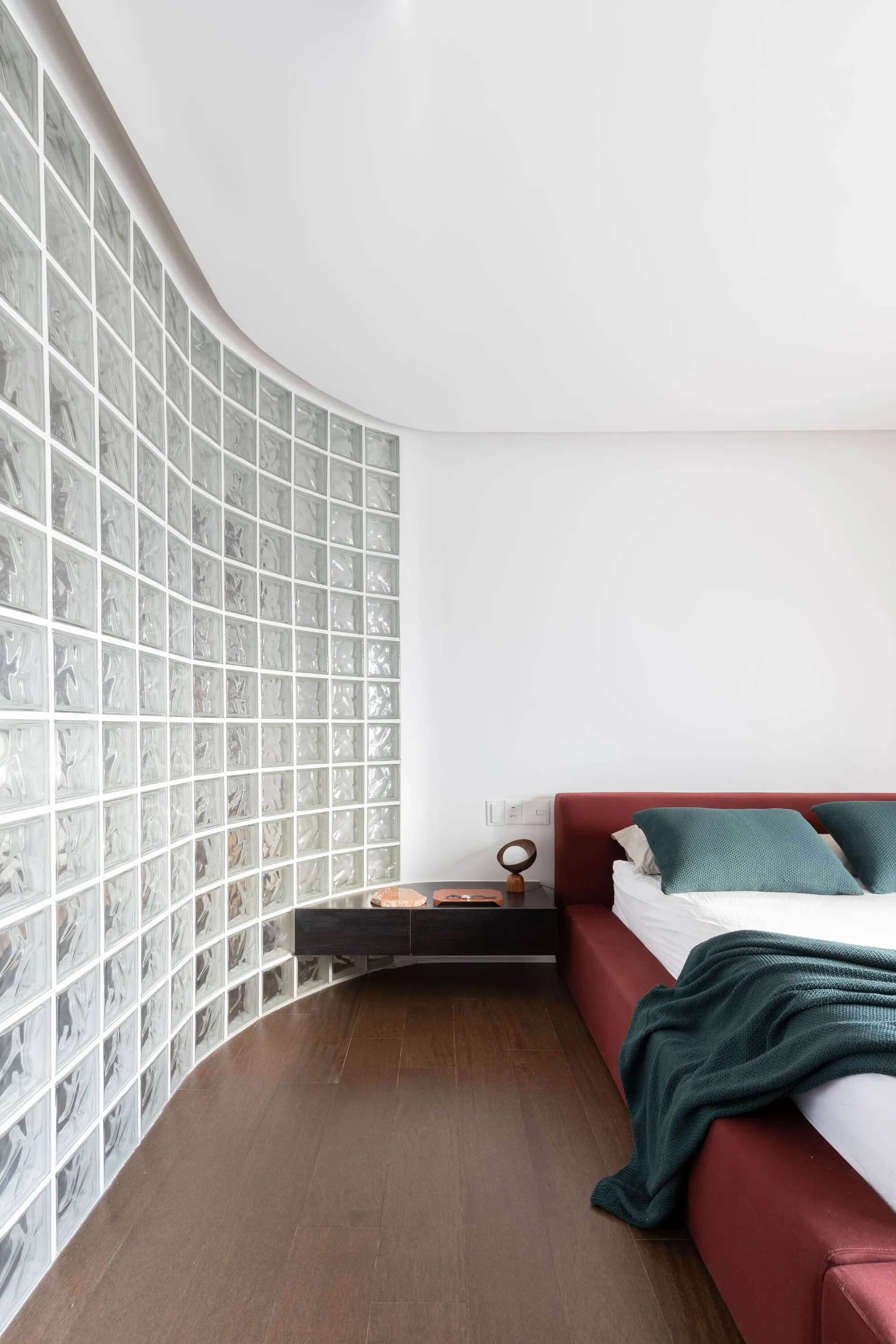 A modern bedroom features a curved glass block partition wall next to a dark wood floating nightstand and a deep red bed with teal pillows.