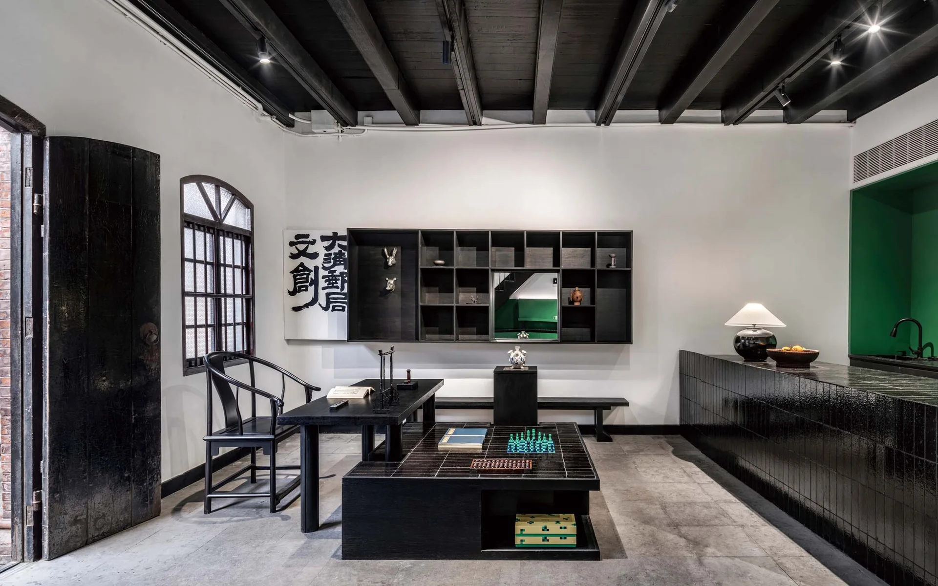 A minimalist study features a black exposed wood beam ceiling, white walls, black wooden furniture, and a bright green accent alcove.