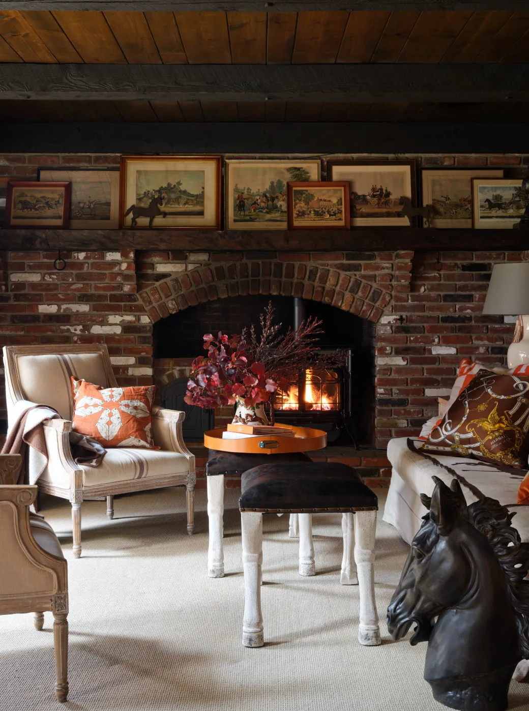A cozy living room features a brick fireplace with framed artwork lined up on the mantel, flanked by a classic armchair and leather stools.