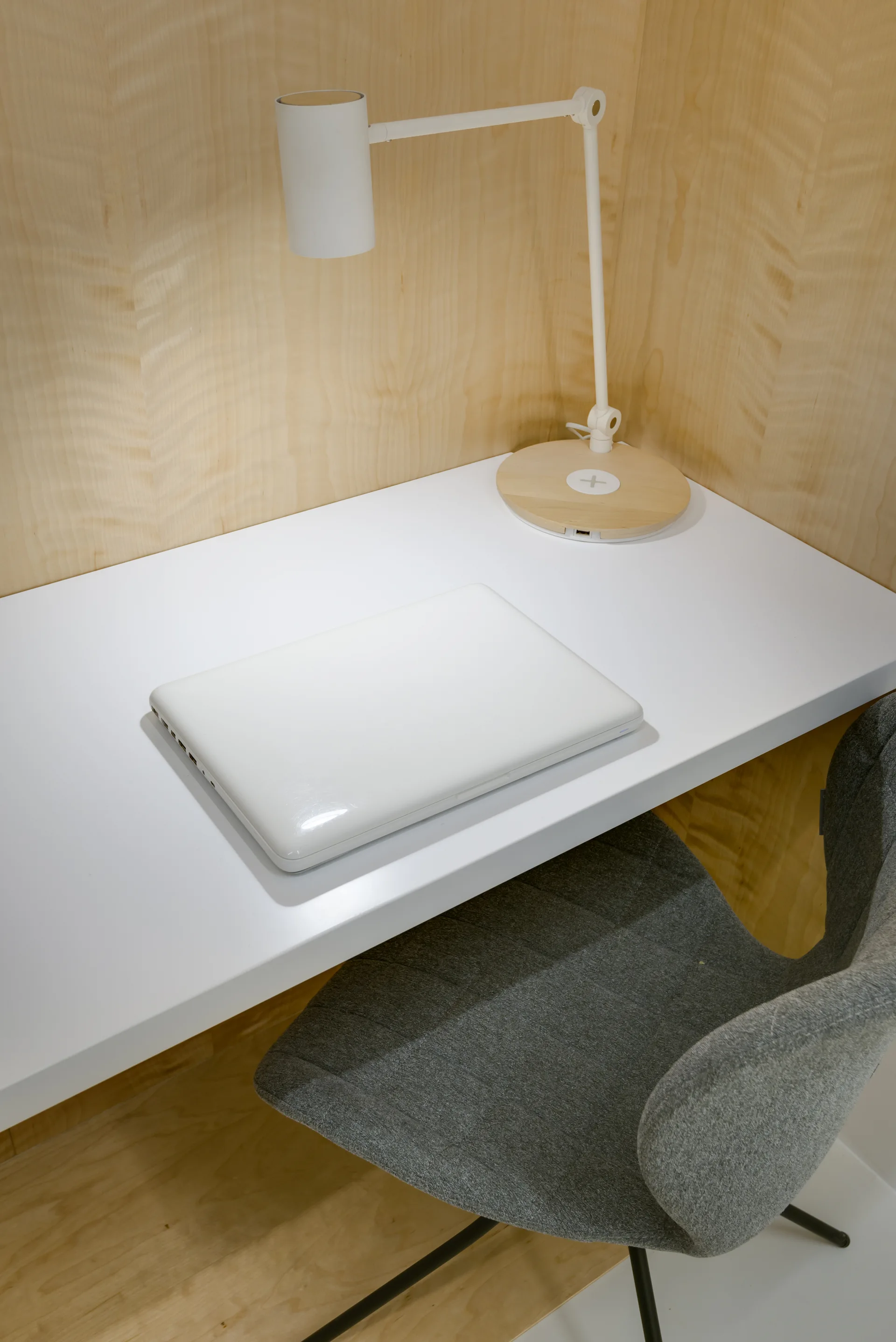 A minimalist office nook with light wood plywood walls features a white desk, grey fabric chair, and a white task lamp.