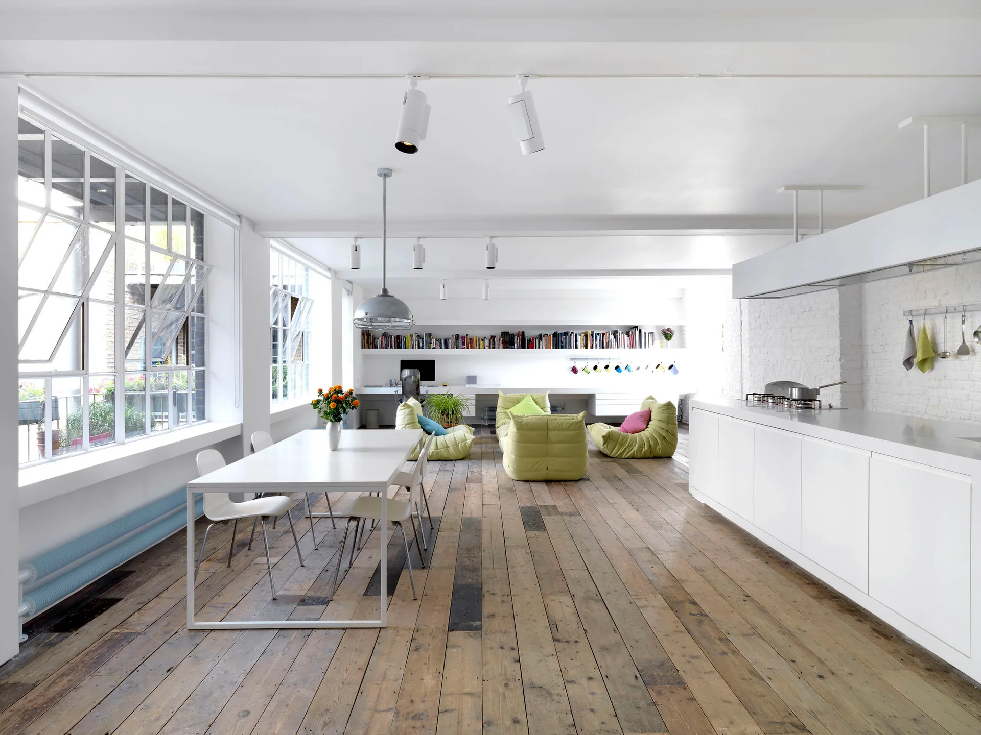 An open-plan white loft features a minimalist kitchen island, a dining table, and green modular chairs over weathered wide-plank wood flooring.