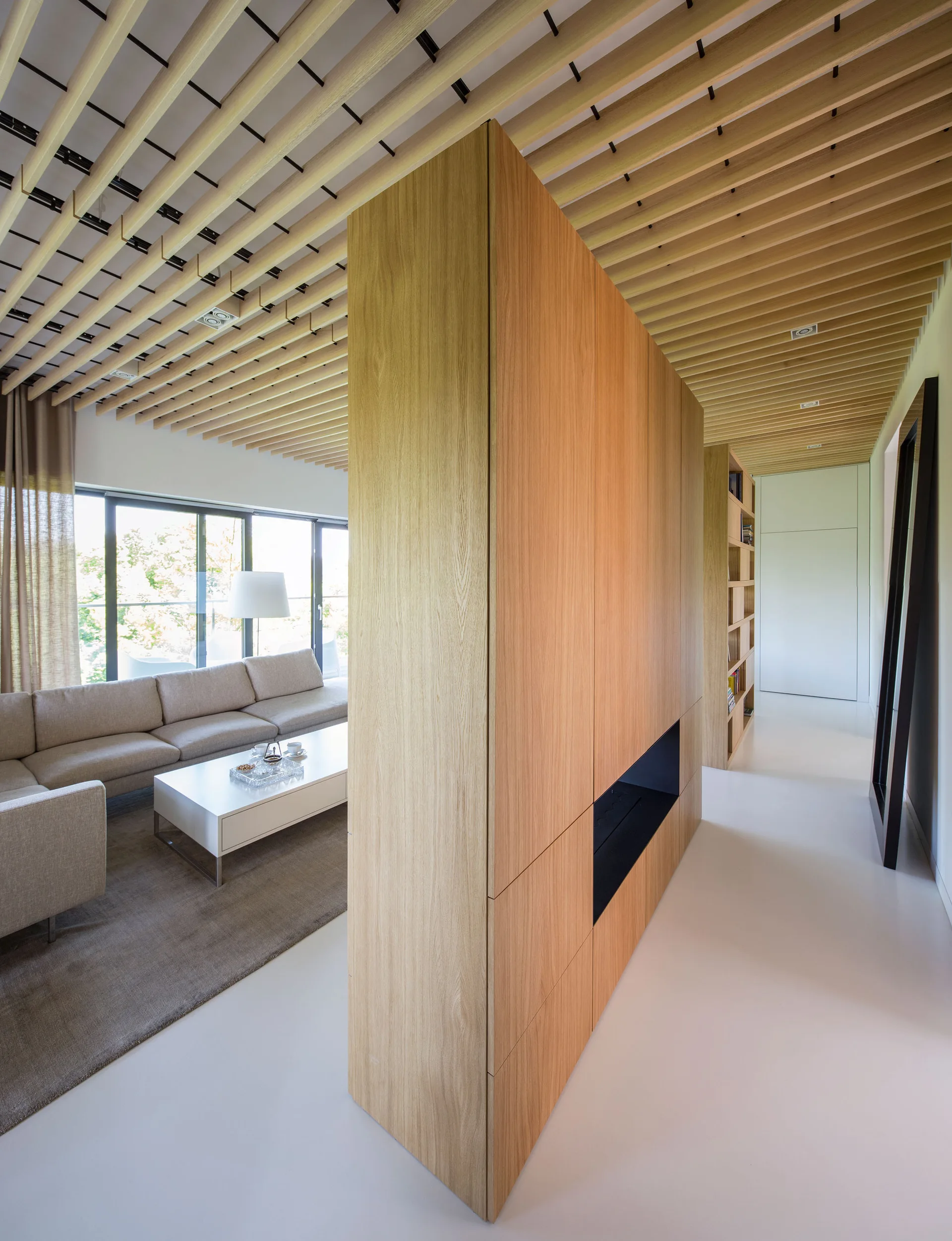 A minimalist living room featuring a light wood room divider with a built-in fireplace beneath a continuous slatted wood ceiling.