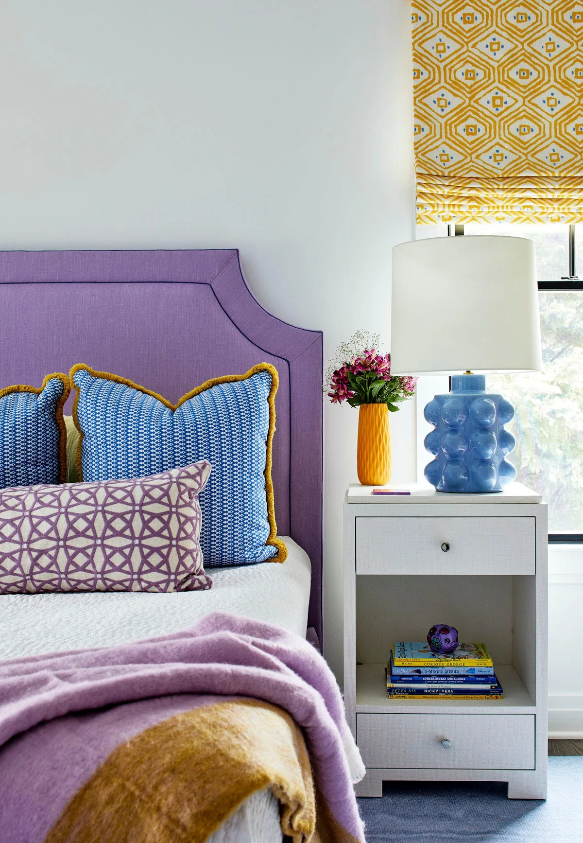 A bedroom corner features a purple upholstered headboard, blue geometric pillows, and a white nightstand holding a blue knobby ceramic lamp.