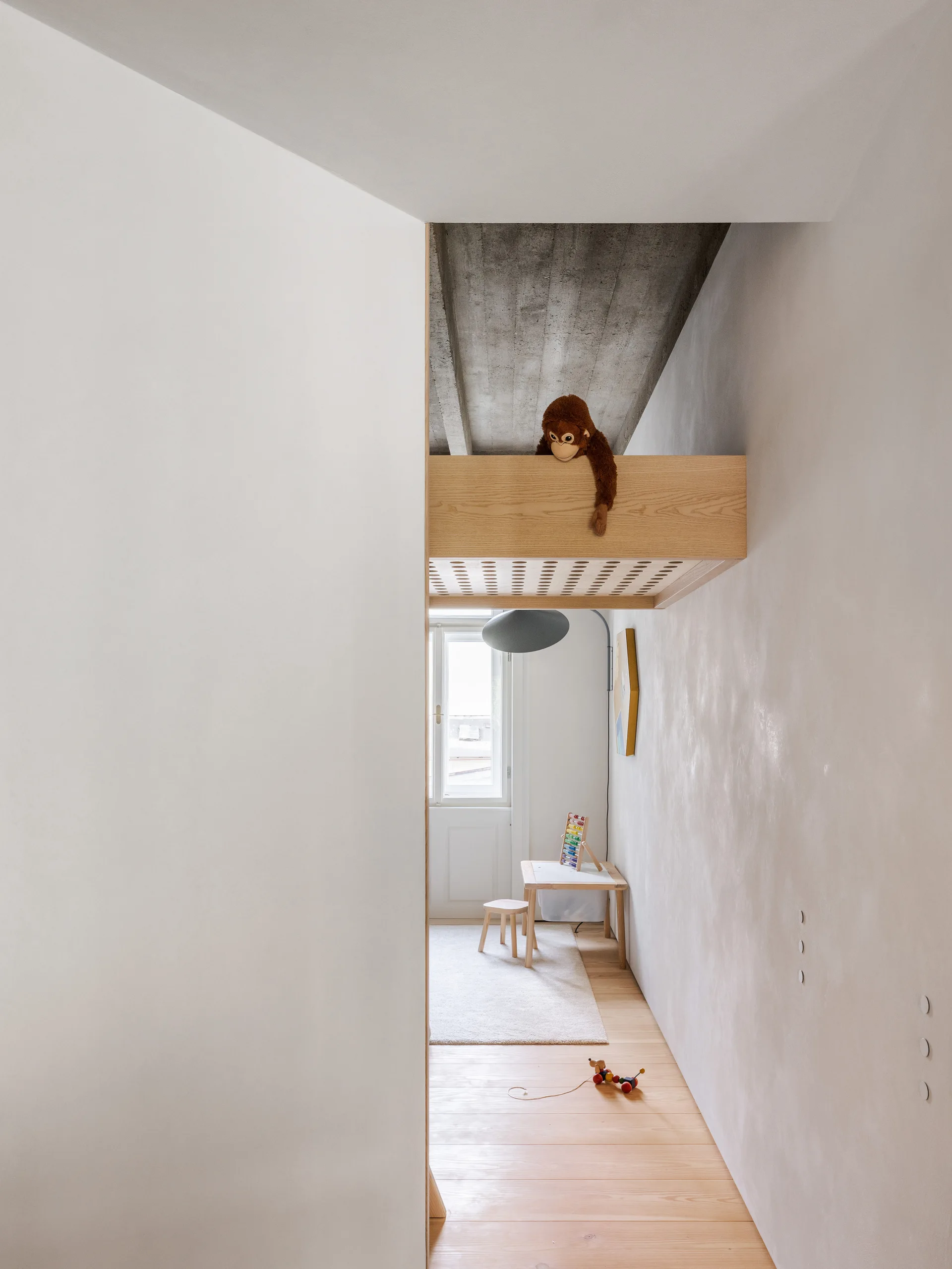 A minimalist kids' room features a light wood loft bed with a plush monkey, a small play table, and a raw concrete ceiling.
