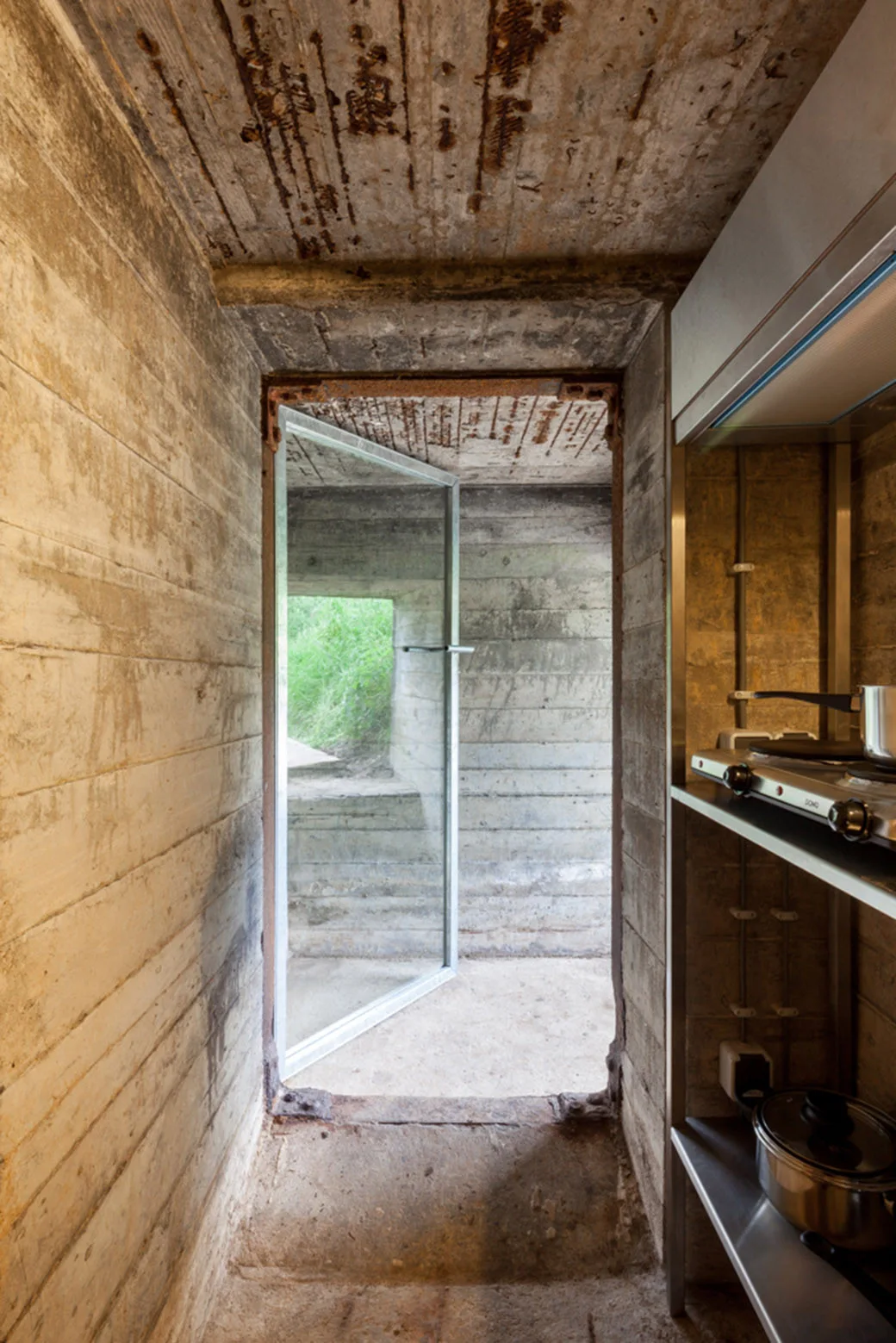 A converted concrete bunker interior featuring board-formed concrete walls, an open metal-framed glass door, and industrial stainless steel kitchen shelving.