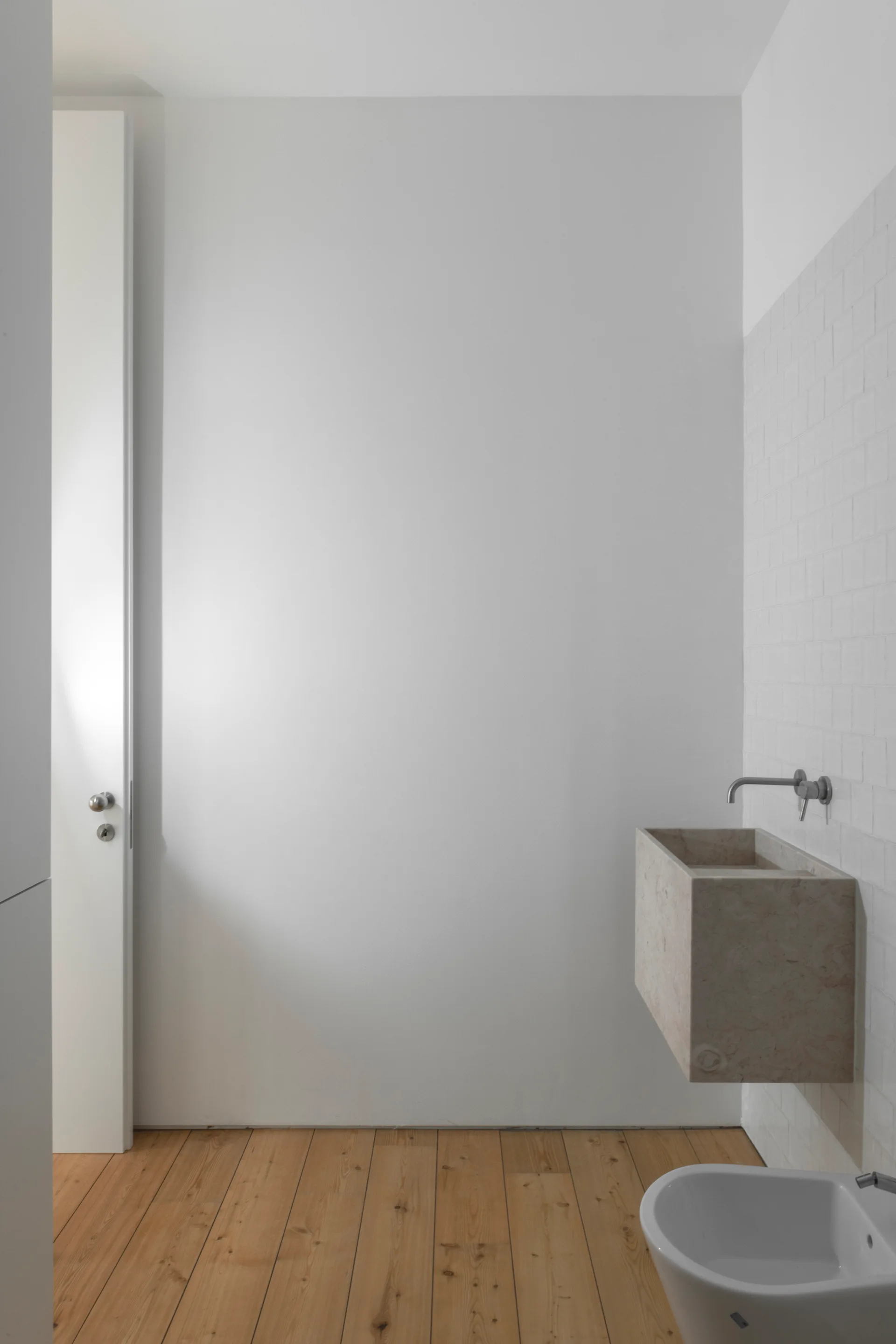 A minimalist bathroom features a wall-mounted rectangular stone sink, a white bidet, white grid wall tiles, and natural wood plank flooring.