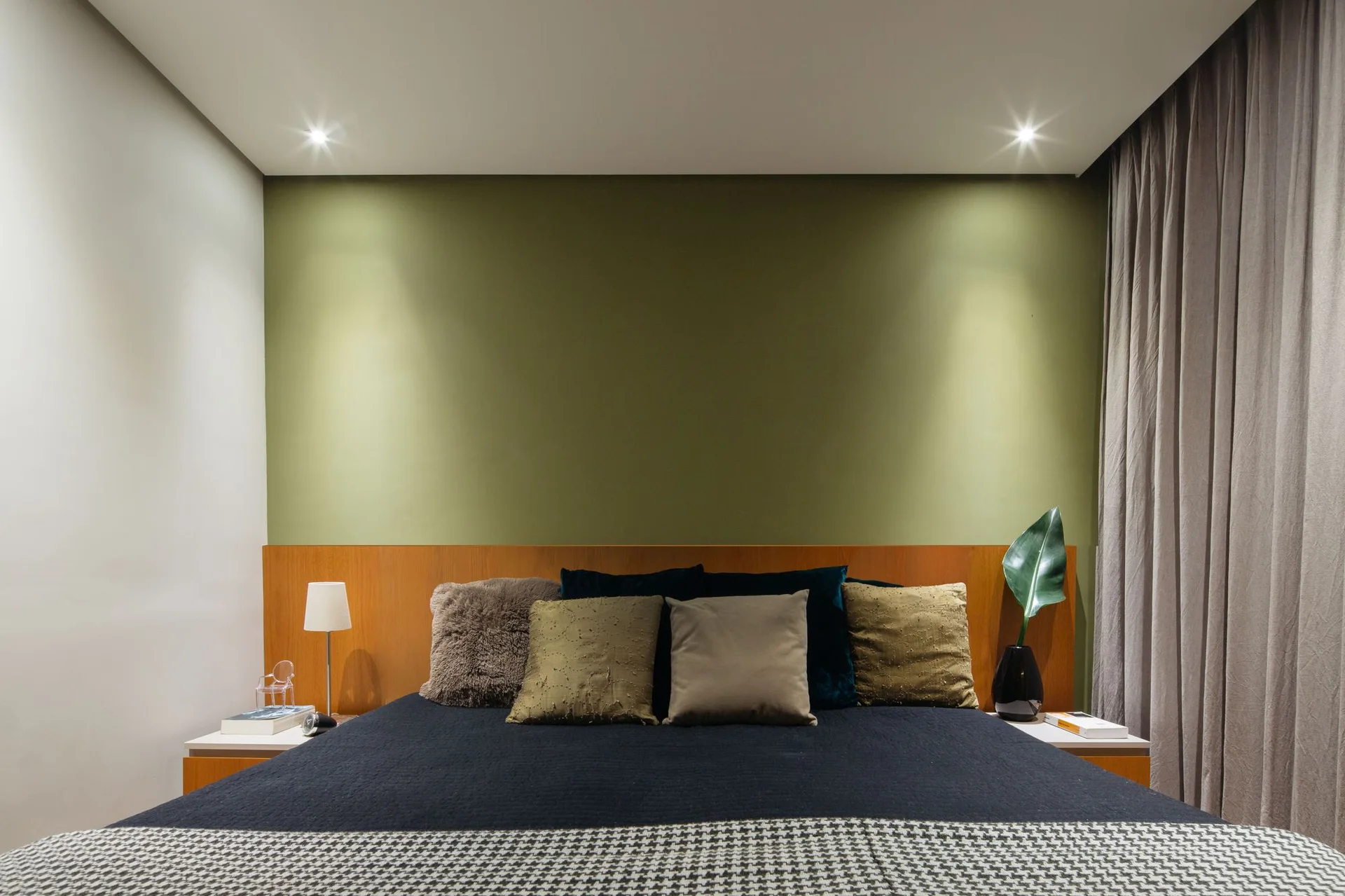 A modern bedroom featuring a bed with patterned pillows and a wide wood-toned headboard against a solid olive green accent wall.