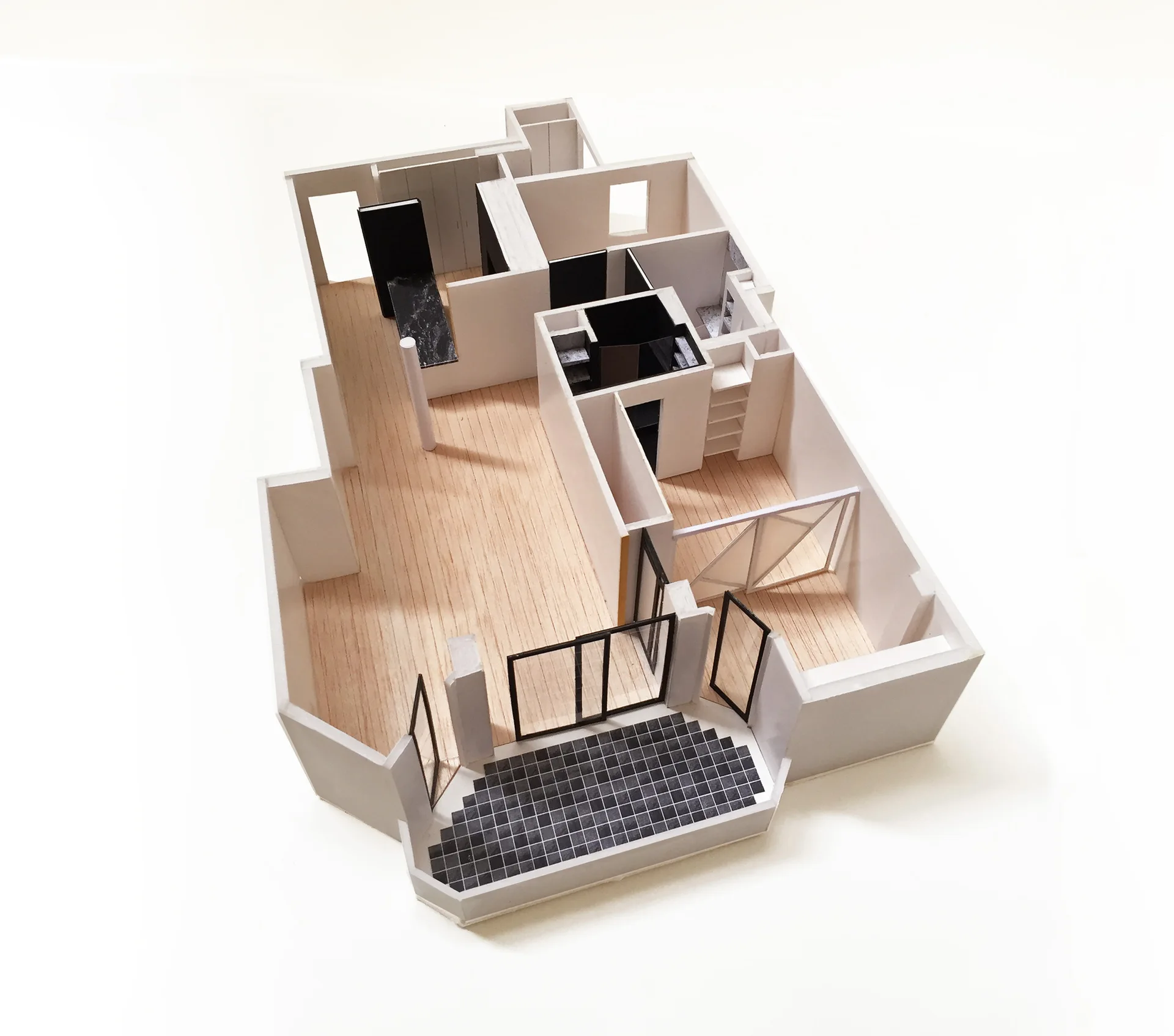 A physical architectural scale model of an apartment floor plan featuring white walls, simulated wood flooring, and a black tiled entrance grid.