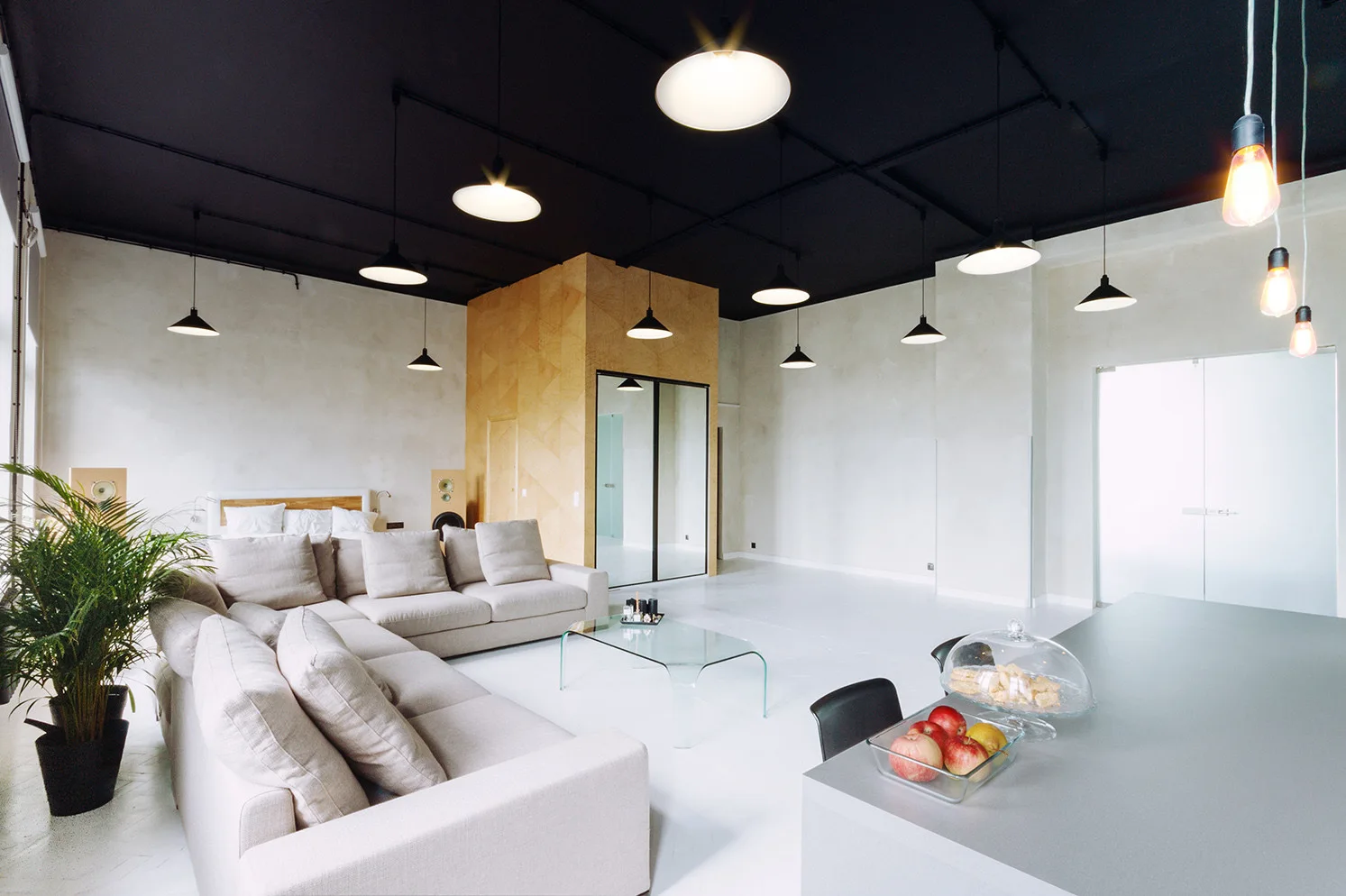 A spacious loft with a black ceiling, modular beige sofa, industrial pendant lights, and a central geometric wood-paneled structure.