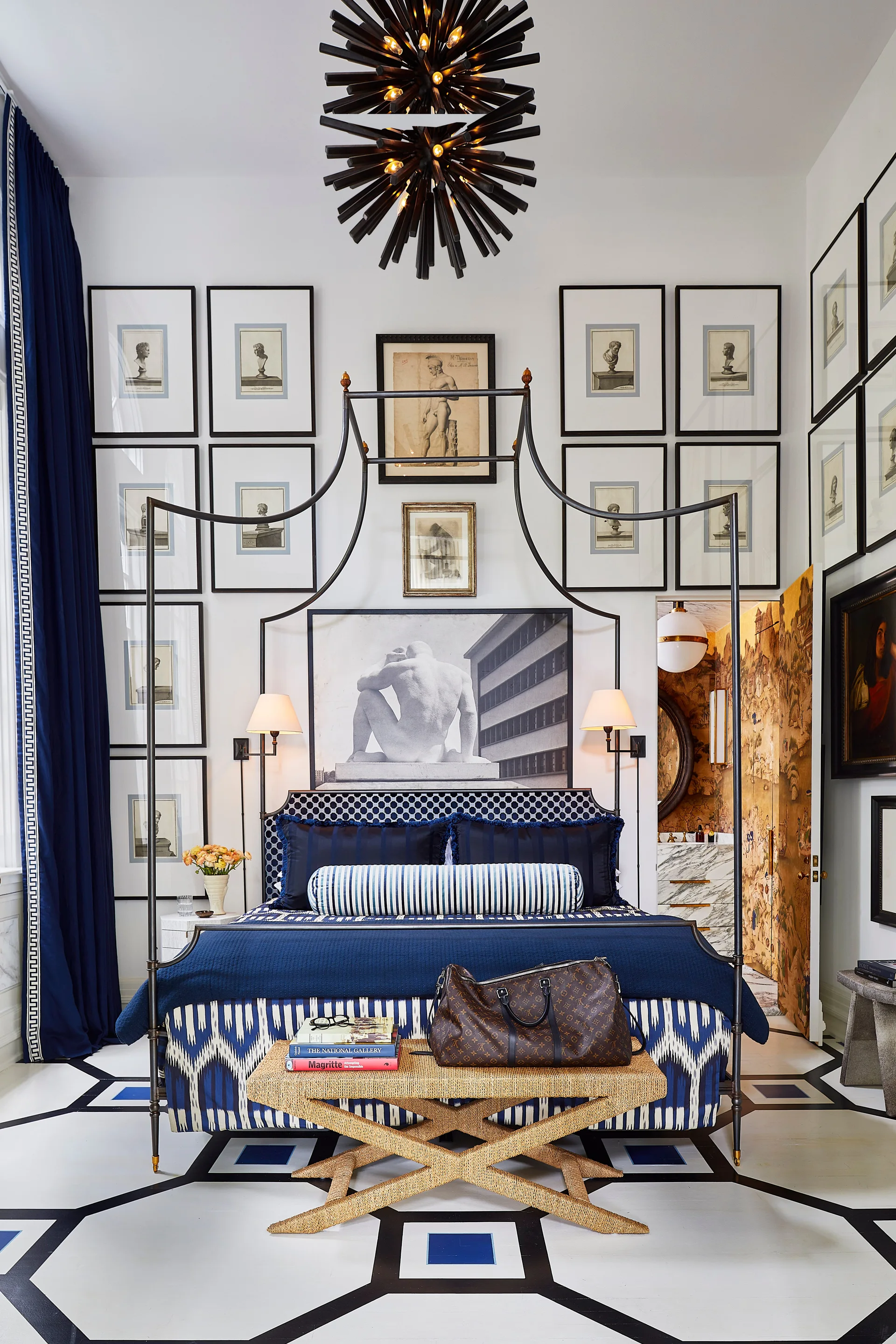 A bedroom features a dark metal canopy bed with blue and white patterned bedding, positioned beneath a large symmetrical grid of framed artwork.