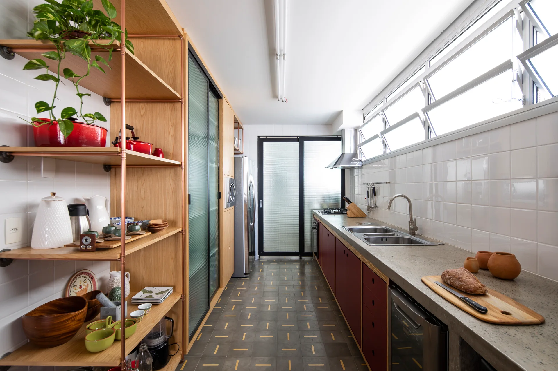 A galley kitchen featuring burgundy base cabinets, concrete countertops, white square-tiled walls, and custom wood shelving with copper pipe supports.