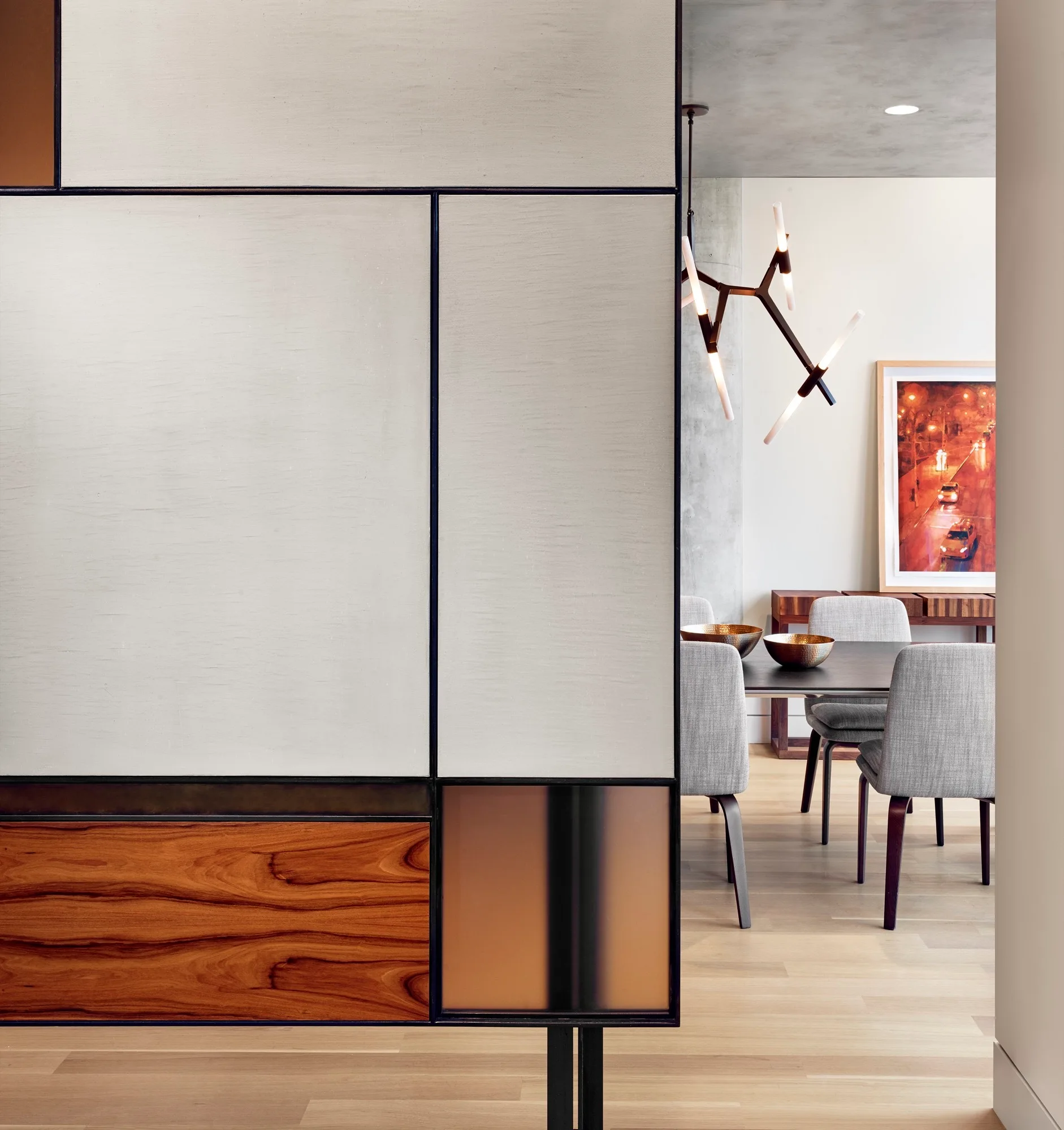 A geometric room divider with wood and glass panels frames a dining area with gray chairs, a dark table, and a branching light fixture.