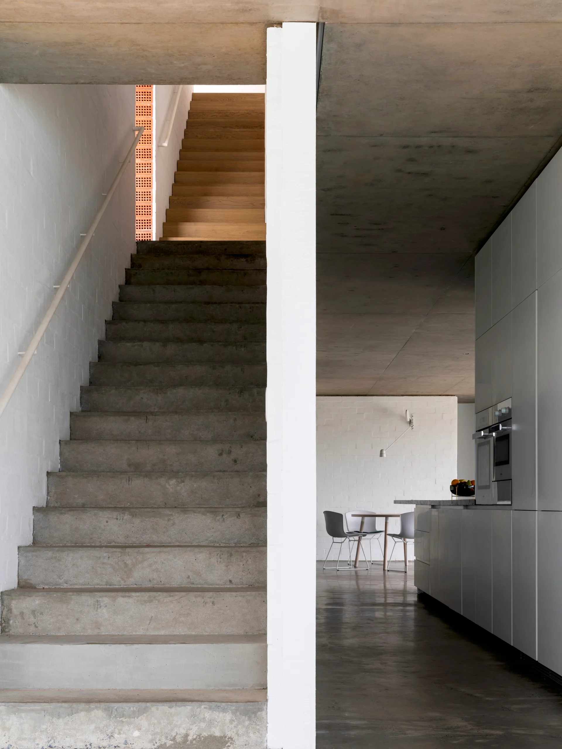 A raw concrete staircase stands next to a modern kitchen with flat white cabinets, polished concrete flooring, and an exposed concrete ceiling.